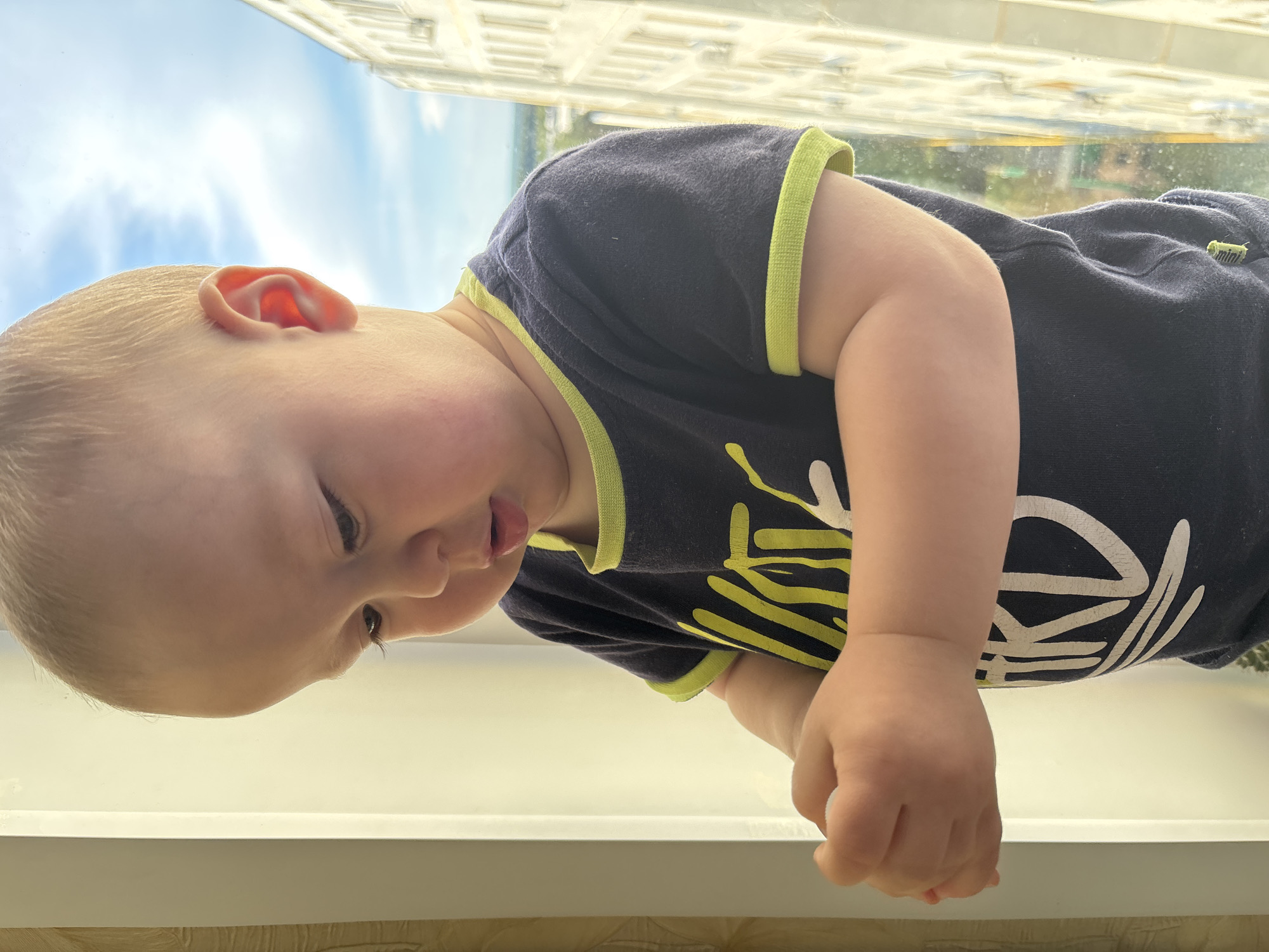 A close-up, head-and-shoulders shot shows a young Caucasian toddler with blond hair, looking downwards at their hands. The toddler is wearing a dark blue t-shirt with a lime green trim around the neckline and sleeves. The t-shirt has yellow and white lettering on it. The toddler is positioned near a window, with a cream-colored vertical frame to their left. Outside the window, a bright, cloudy sky is visible, along with a tall building with many windows, characteristic of a residential building in the city of Naberezhnye Chelny, Russia. The lighting suggests it is daytime, possibly late afternoon, due to the warm glow. The toddler's hands are held together, and they appear to be holding something small and white. The background shows the urban landscape.