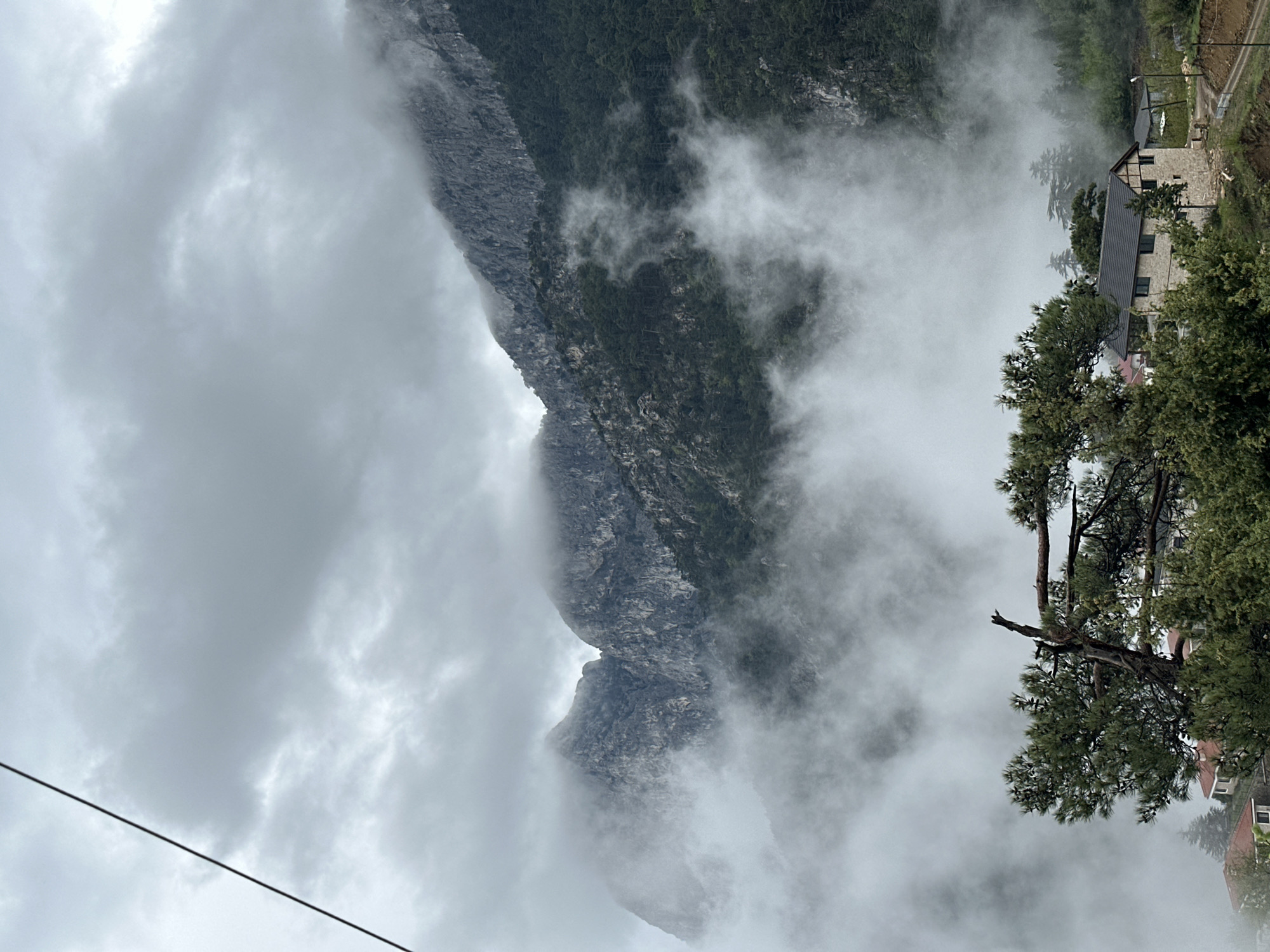 The image depicts a mountainous landscape shrouded in fog, likely located near a residential area. A steep, rocky mountain face dominates the left side of the frame. The fog obscures much of the mountain's details, but the texture of the rock is still discernible. On the right side of the image, there are trees and buildings. The architecture appears to be European in style. An electrical wire is visible in the lower-left corner of the frame. The sky is overcast, suggesting a cloudy or rainy day. The location is consistent with the mountainous terrain near Konyaaltı, Turkey, where residential areas are situated near the mountains.
