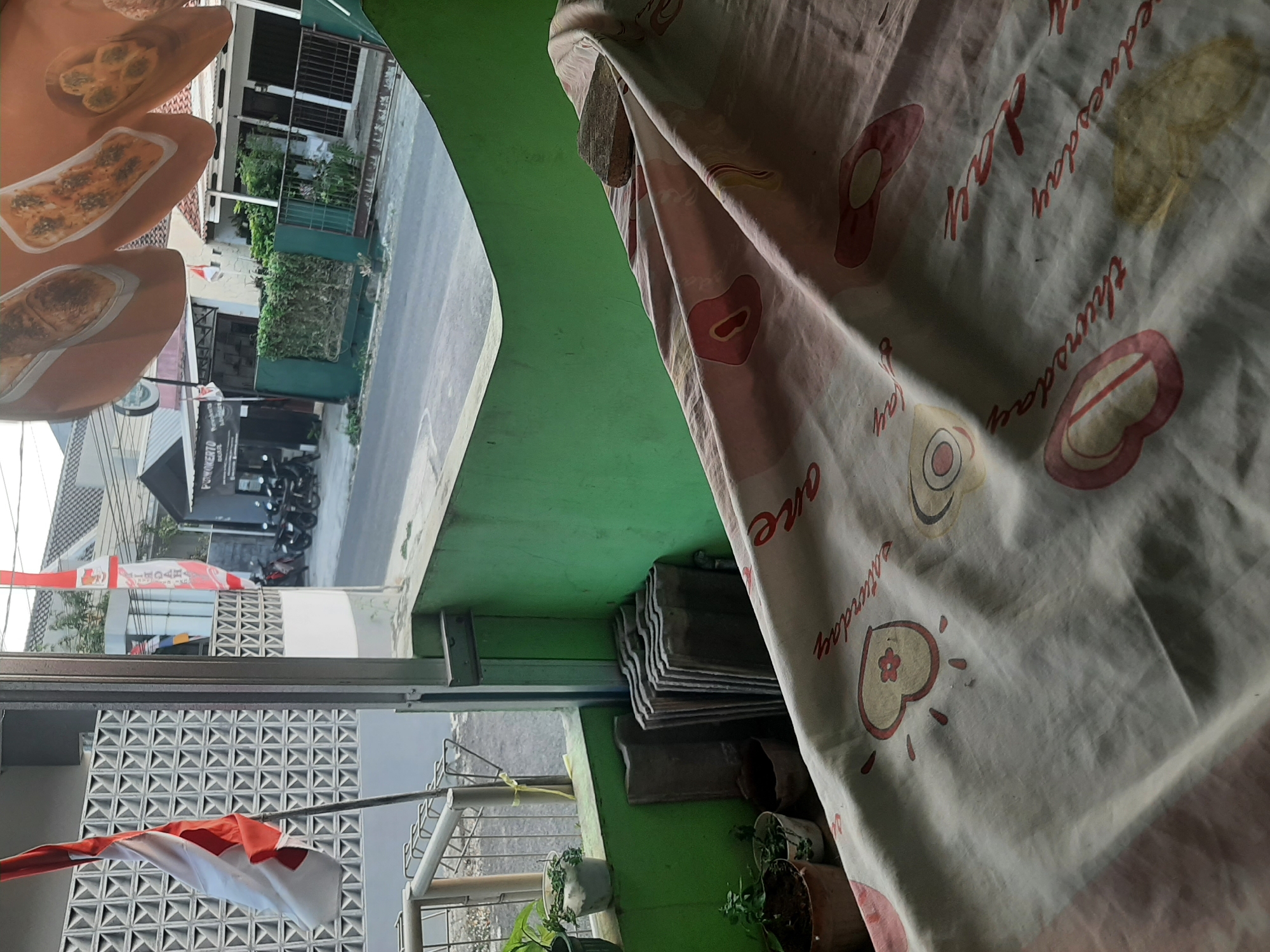 The image depicts a street view from a covered outdoor area in Sumampir, Indonesia. The foreground features a patterned fabric draped over a surface, with a stone resting on it. To the left, a red and white Indonesian flag is visible, attached to a flagpole and fluttering slightly. Behind the flag, a wall with a geometric pattern is present, along with several potted plants and stacks of roof tiles. The middle ground shows a street with parked motorcycles and a building with the signage "POM KEPTO" in red lettering. The background reveals residential buildings and lush greenery. The overall scene suggests a residential or commercial street in a tropical climate.