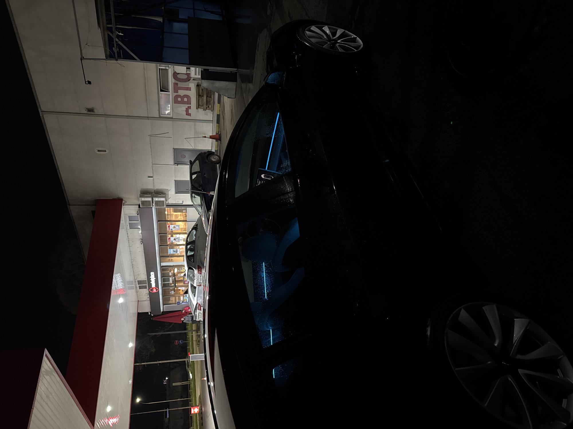 A nighttime view in Varna, Bulgaria, captures a car, possibly a Tesla due to its modern wheel design and interior blue lighting, parked near a gas station. The gas station has red and white branding, and a convenience store is visible in the background, with cars parked alongside. In the distance, signage with Cyrillic script is present. The environment is dark with streetlights and gas station lights providing the only illumination.