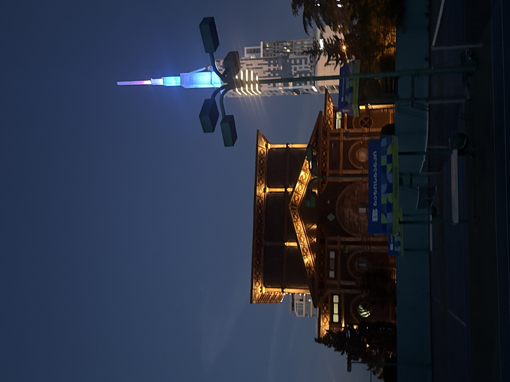 The image shows a nighttime scene in Batumi, Georgia, featuring the Astronomical Clock Tower and a nearby futuristic light structure. The clock tower, lit with golden lights, has an ornate design and sits near modern high-rise buildings. A blue and purple-colored, spire-like structure is also visible in the background. The area appears to be a public space, indicated by the presence of what seems to be a tennis court and other urban fixtures. The scene is at dusk or night, given the deep blue sky.