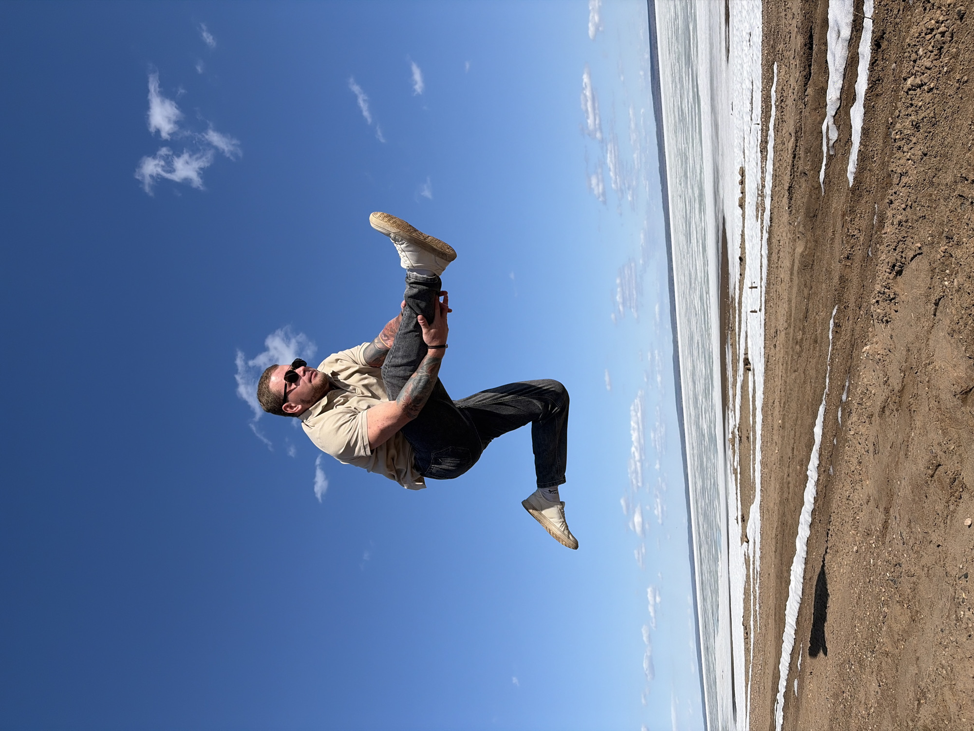 A male individual wearing sunglasses, a beige shirt, dark jeans, and white sneakers is upside down in the air against a bright blue sky with scattered white clouds. His right arm is bent and holding his right leg up, and his left arm is bent across his chest. He appears to be tattooed on his left arm. The background shows a thin strip of what looks like a frozen body of water next to a sandy beach with patches of snow or ice. The photo was likely taken in Chaikovsky, Russia, given the prompt.