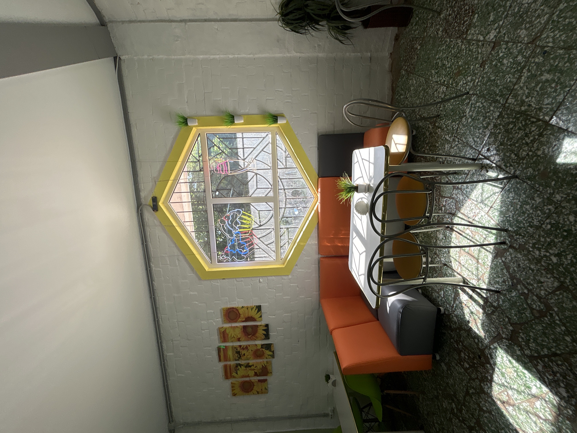 This is an indoor scene of a cafe. A hexagonal window with yellow trim is a central feature, revealing a view of trees and sunlight outside. The window has decorative stained glass depicting ice cream cones. Inside, an orange upholstered booth seating area is arranged around a white-topped table with chrome legs. Two chairs with yellow seats and chrome frames are pulled up to the table. A plant in a white pot sits on the windowsill, and another small plant is on the table. The walls are white brick, and the floor is a speckled green and white terrazzo. The scene is illuminated by bright sunlight casting distinct shadows on the floor. There are no people visible in the image. Based on the limited visual information, it is not possible to definitively identify the specific city or country, and therefore the location in Rodniki, Russia cannot be confirmed.