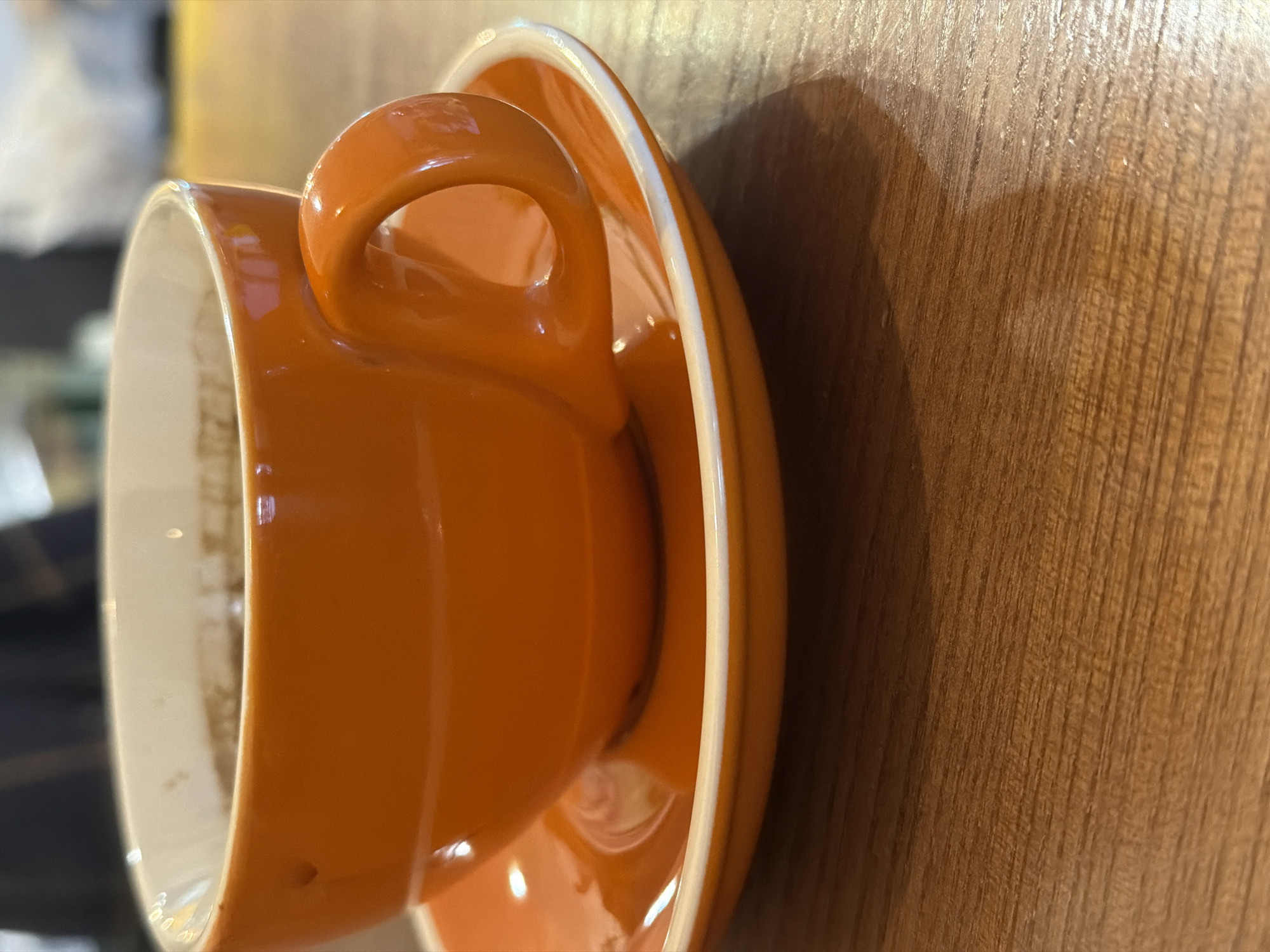 An orange cup and saucer are seen in close-up, placed on a wooden surface. The cup is predominantly orange with a white interior lining. The saucer complements the cup in color and design. The scene appears to be indoors. The presence of the cup and saucer suggests a possible setting like a cafe or a kitchen. The location in the city of Dnipro, Ukraine is not discernible from the visual information provided.