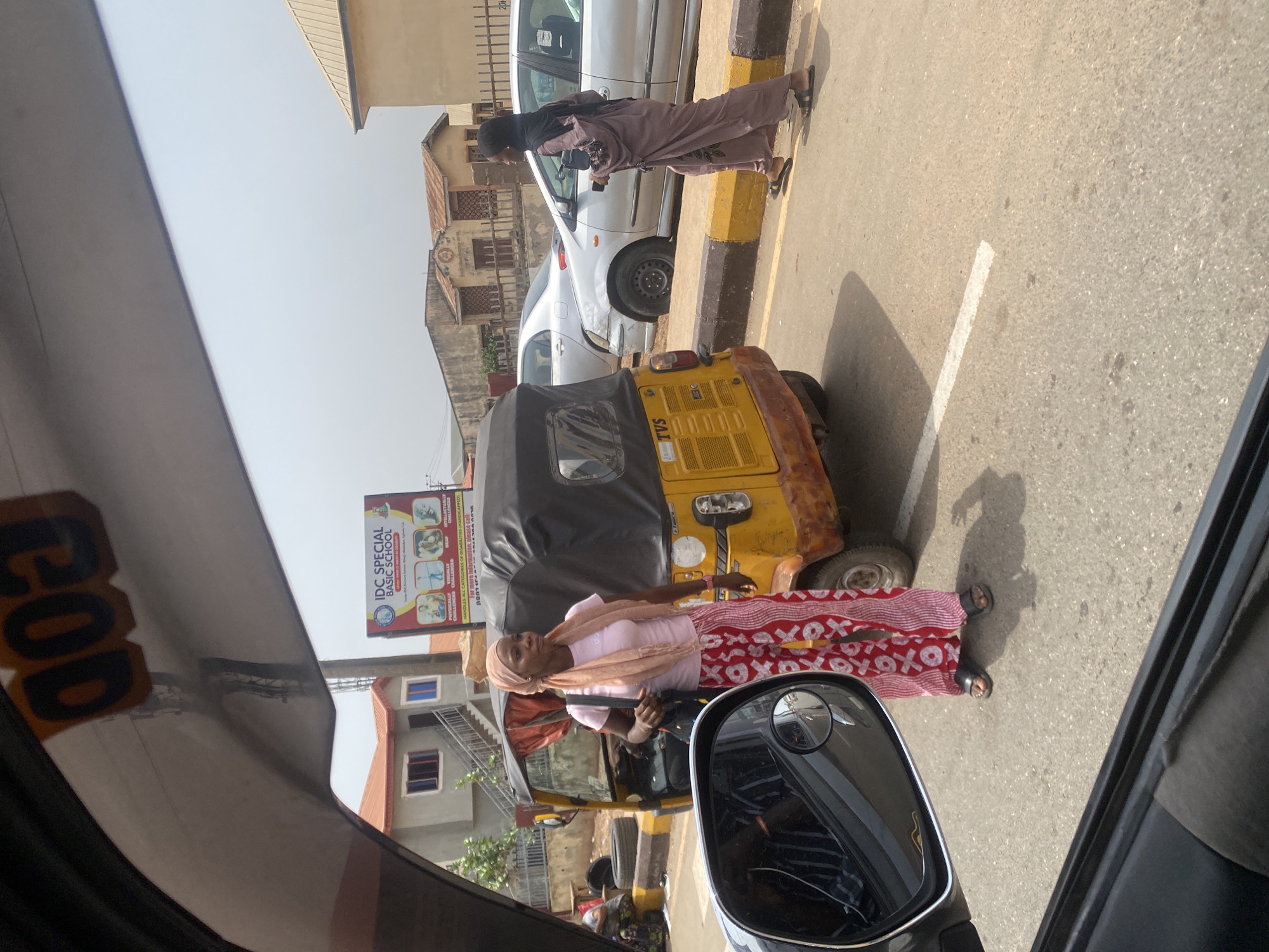 The image shows a street scene in AlakIa, Nigeria. A woman wearing a headscarf, light pink shirt, and red and white patterned skirt stands next to a yellow auto-rickshaw (TVS brand). Another woman in a hijab is walking along the road next to a white car. A sign for "IDC Special Basic School" is visible in the background. The view is taken from inside a vehicle, with parts of the dashboard and side mirror visible.