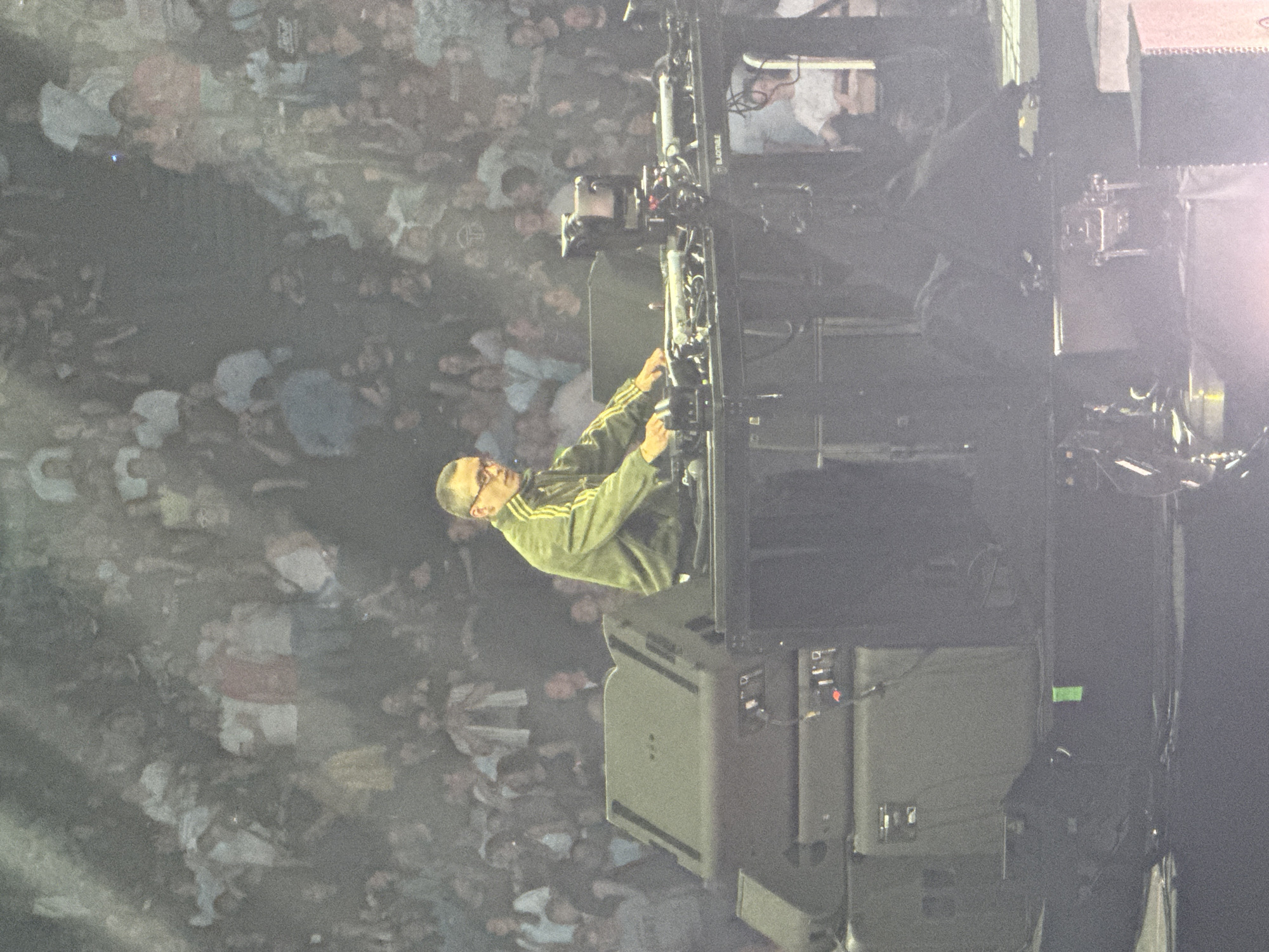 A wide-angle photograph shows a person in a green track suit standing next to a sound speaker system. They are wearing glasses and appear to be making adjustments to the equipment. The background is a crowd of people. The photograph appears to be taken from an elevated position. The location is not identifiable as Paris, France.