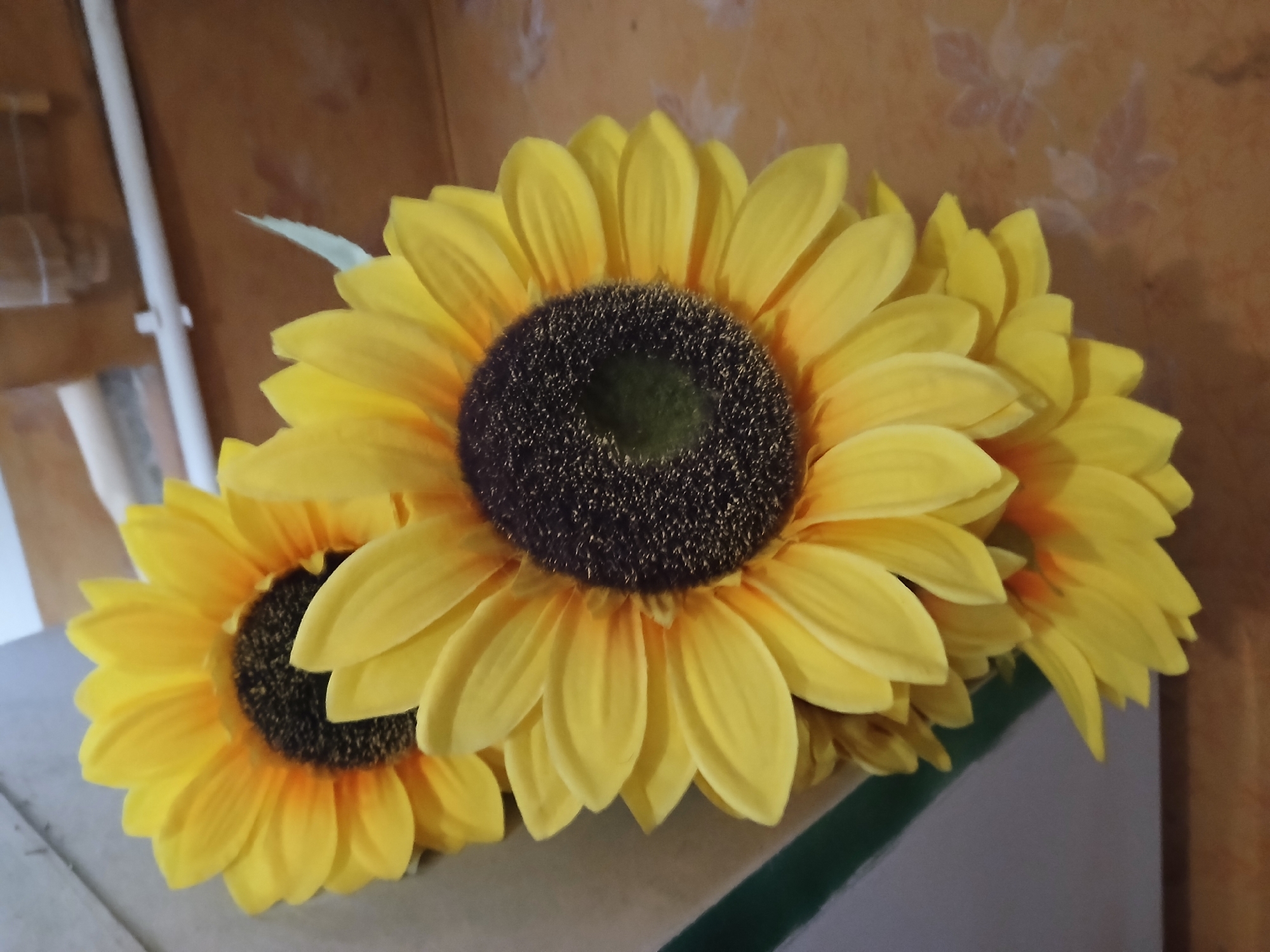 The image shows a close-up view of three artificial sunflowers arranged together. The sunflowers have bright yellow petals and dark brown centers with a hint of green in the very middle. The largest sunflower is in the foreground, facing slightly to the right, while two smaller ones are positioned to its left and behind. The background is a warm-toned, textured wall that appears to have a faint floral pattern. A white pipe or rod is visible on the far left, running vertically. A shelf or surface is partially visible in the lower portion of the image, with a dark green strip along its edge. The lighting is warm and appears to come from the right, casting subtle shadows. There are no people or animals present in the image. The scene is indoors, likely in a domestic setting. There is no information in the image to suggest a specific location within the city of Lipetsk, Russia.