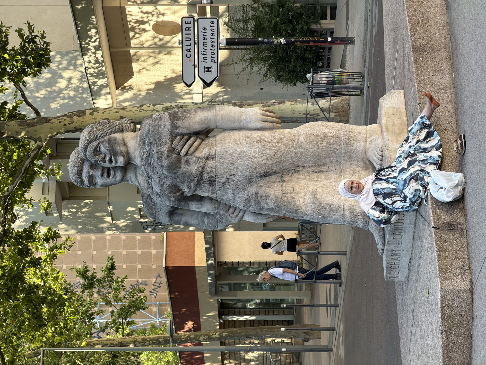 The image depicts a large stone sculpture of a couple embracing, positioned on a concrete pedestal in an outdoor urban setting. The sculpture's base is inscribed with the words "LE CHANT des CANUTS." To the right of the sculpture, a street sign on a pole indicates directions to "CALUIRE" and "infirmerie protestante."
In the foreground, a woman wearing a light pink hijab and a patterned dress is seated on the edge of the concrete pedestal, facing the viewer and slightly to the right. Her left leg is extended, and she is wearing sandals. A white plastic bag is next to her left.
In the middle ground, behind the sculpture and to the left, a man with graying hair, wearing a white polo shirt and dark pants, walks away from the viewer, carrying a backpack. Further in the background, a woman in a white top and black skirt walks away from the viewer. Beyond them, a building with a red and beige facade is visible. Trees with green foliage are present throughout the scene, casting shadows on the buildings and ground.
The scene appears to be on a sunny day, with natural light illuminating the area. The overall impression is a public space in a city, likely Lyon, France, given the presence of the street signs and the inscription "CANUTS," which refers to the silk weavers of Lyon.