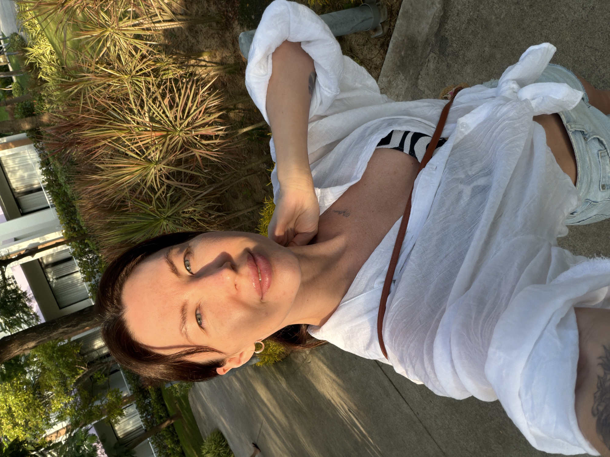 In Dong Ninh Hoa Ward, Vietnam, a woman with dark hair stands outdoors on a paved path, possibly a sidewalk, at golden hour. She is wearing a white, open-collared, long-sleeved shirt, tied at the waist, revealing a black and white striped bikini top and light-wash denim shorts. She has a brown crossbody bag strap visible over her shoulder. Her left arm is raised, touching her neck, and she is looking towards the camera. The background features lush green trees and foliage, with a modern building with light-colored walls and windows visible behind the greenery. The sunlight casts long shadows across the path and illuminates the scene with a warm glow.