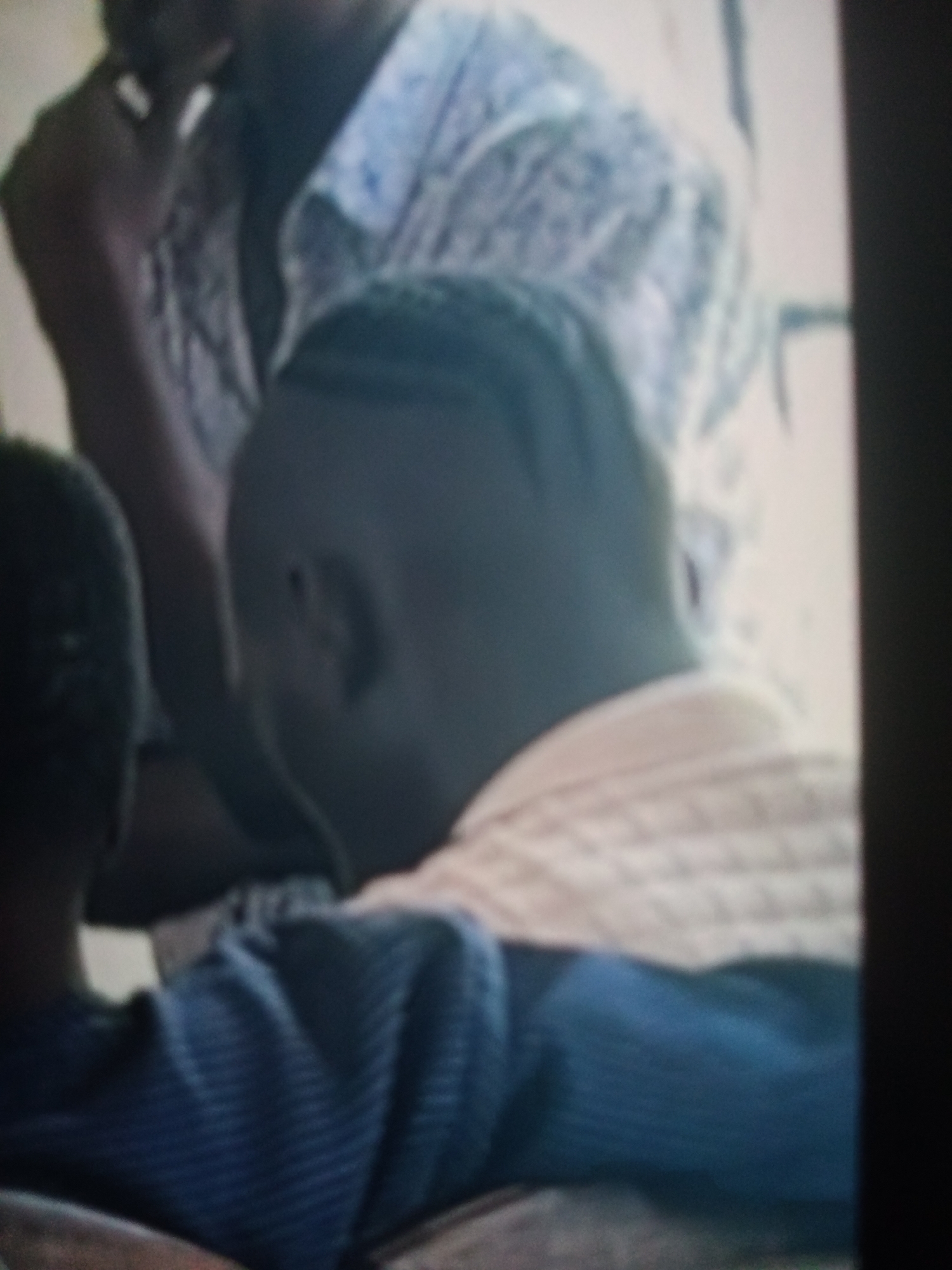 The image depicts an indoor scene with at least three individuals partially visible. In the foreground, a person wearing a dark blue, ribbed garment is seated, facing away from the camera. To their right, another individual's head and shoulders are visible, wearing a light-colored, textured garment. Further back, a third person is seen in profile, with their arm resting on their head. They are wearing a blue and white patterned shirt. The environment appears to be a room with plain walls, suggesting it could be a school, community center, or residential area in Jalingo, Nigeria. The lighting is soft, and the image quality is low, indicating it may be an older photograph or a low-resolution capture. There is no specific detail to pinpoint the exact location within Jalingo, but the context suggests a setting where people are gathered, possibly for a meeting, class, or social event.