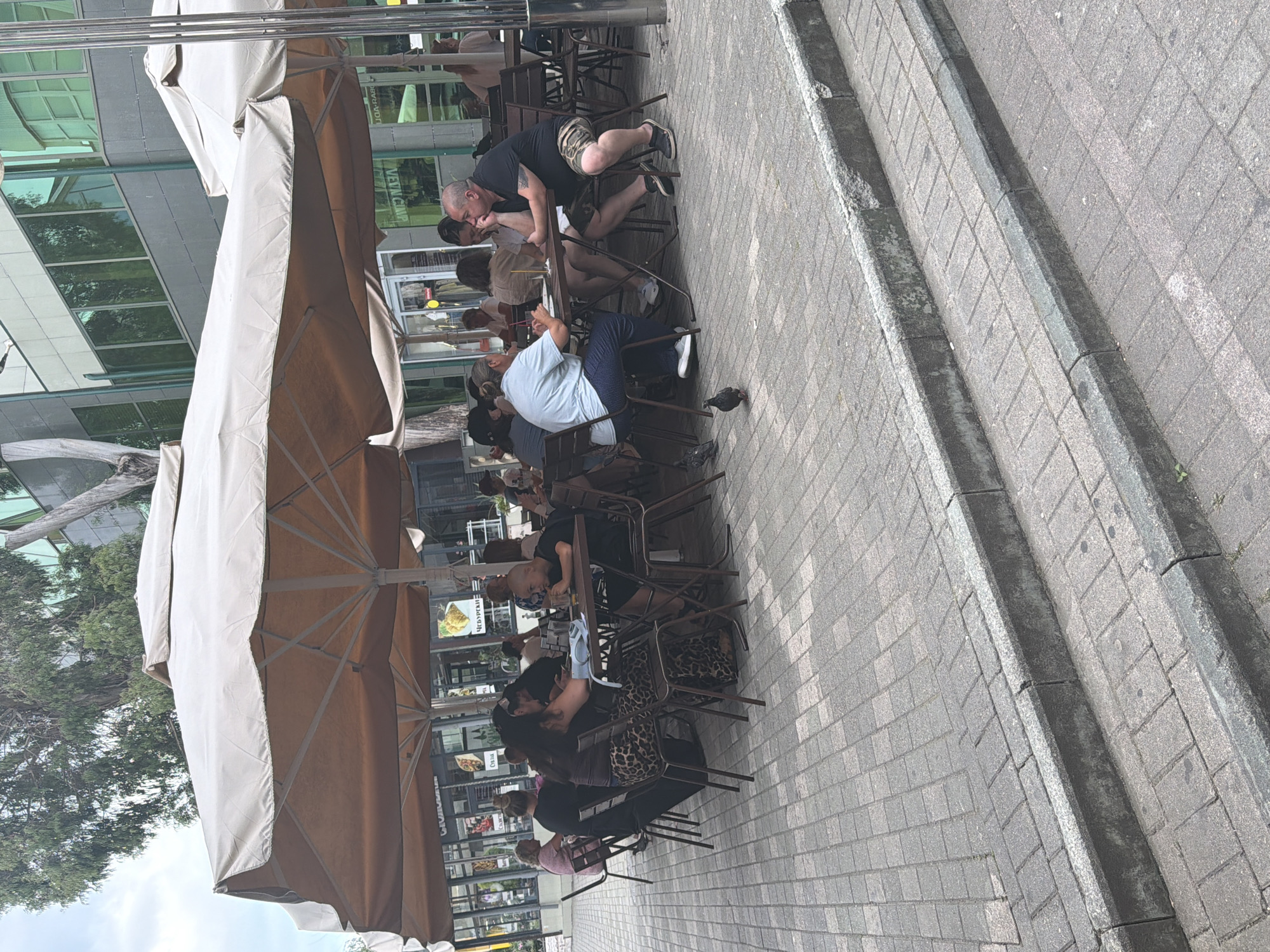 The image depicts an outdoor seating area of a cafe, with several tables and chairs beneath large, beige umbrellas. People are seated at the tables, engaged in various activities such as eating, drinking, and conversing. Some individuals appear to be in close conversation, while others are more casually observing their surroundings.
In the foreground, a man wearing a black t-shirt and camouflage shorts is leaning over a table, seemingly engaged in an intimate moment with another person. To his left, a person in a light blue shirt sits at a table, looking down. Further back, more patrons are visible, some with their faces turned away from the camera. The cafe is situated on a paved area with cobblestone patterns. A narrow, dark channel runs along the side of the paved area, separating it from the sidewalk.
In the background, a modern building with large glass windows reflects the surrounding environment, including trees. The time of day appears to be daytime, and the weather seems pleasant, as people are comfortably seated outdoors. The overall atmosphere suggests a relaxed social gathering.
There is some visible text on signs in the background, including what appears to be Russian script on posters or menus displayed within the cafe's windows. For instance, one sign appears to read "ЧЕРНОМОРСКИЙ" and another includes "ВЕНТИЛЯЦИЯ".