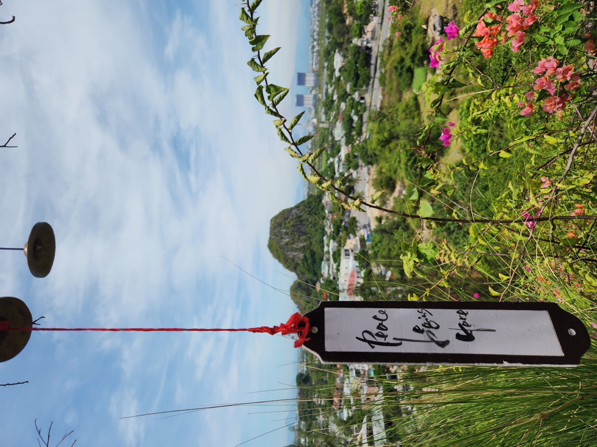 A vertical, close-up shot captures a hanging sign with the words "Peace is here" in elegant calligraphy. The sign, attached to a red string, is positioned in the foreground, slightly to the left. Behind it, a lush green landscape unfolds, dotted with houses and trees, leading to a prominent, rocky hill in the middle ground. In the background, the city of Da Nang, Vietnam, is visible with modern buildings and a hazy sky. The image is dominated by natural elements, with vibrant pink and orange bougainvillea flowers adding pops of color in the lower right corner. The overall mood is serene and tranquil, with the sign serving as a focal point against the scenic backdrop.