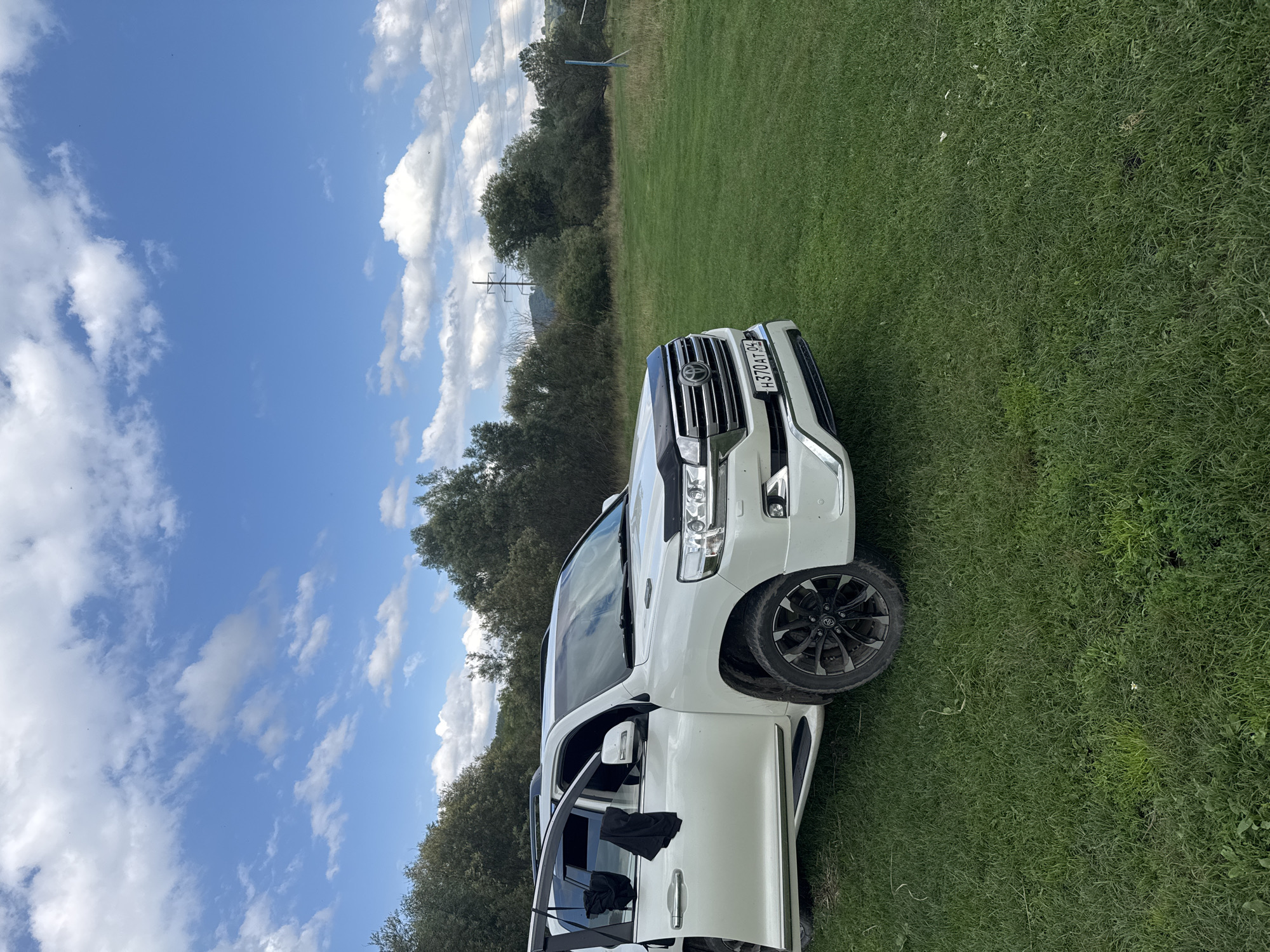 A white Toyota Land Cruiser is parked on a grassy field under a partially cloudy sky. The front passenger door of the vehicle is open, with a black piece of clothing hanging from the interior. The vehicle has a black hood protector and a license plate that reads "H370AT 04". The background consists of a dense line of trees, with a power line tower visible in the distance. The location is the village of Soloneshnoye, Russia, a rural area characterized by natural landscapes.