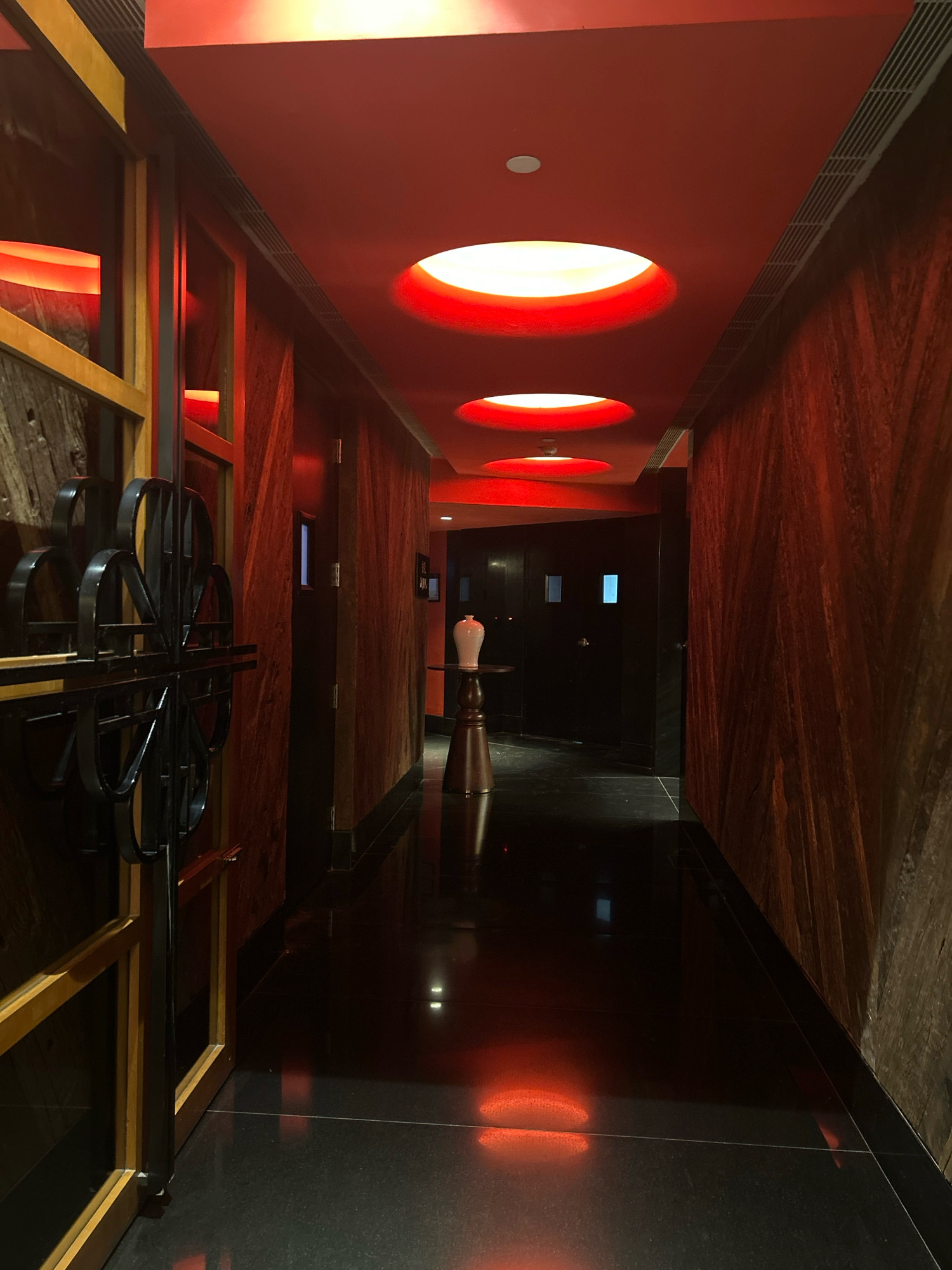 The image shows an interior view of a dimly lit corridor with red overhead lighting. The walls are paneled with dark wood, and the floor is a highly reflective black surface. Several circular recessed lights with red glows are visible in the ceiling. On the left side of the corridor, there is a decorative black metal gate or screen. Further down the corridor, a white vase sits on a dark wooden pedestal. The overall aesthetic is modern and atmospheric. The location is specified as Hyderabad, India.