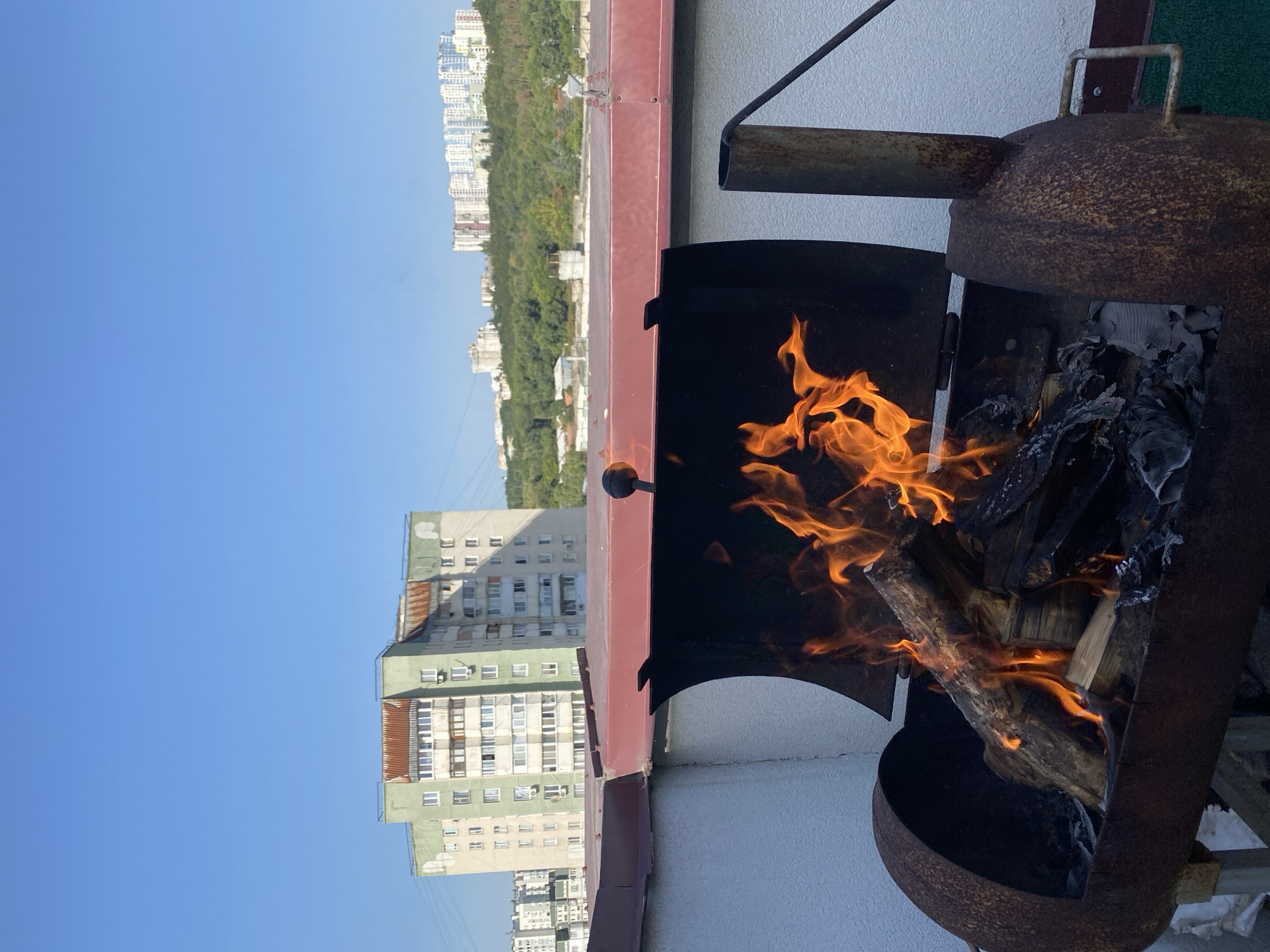The image depicts an outdoor scene on a rooftop with a clear blue sky. In the foreground, a rusty, open barbecue grill contains burning logs and embers. The grill has a curved body and a hinged lid that is partially open. Behind the grill, a red-colored roof structure is visible, extending across the frame. In the background, several multi-story apartment buildings and green, tree-covered hills are seen, suggesting an urban setting. The architecture of the buildings appears consistent with residential areas found in Chișinău, Moldova, characterized by their pastel-colored facades and balconies. The overall scene conveys a sense of leisure and outdoor cooking.