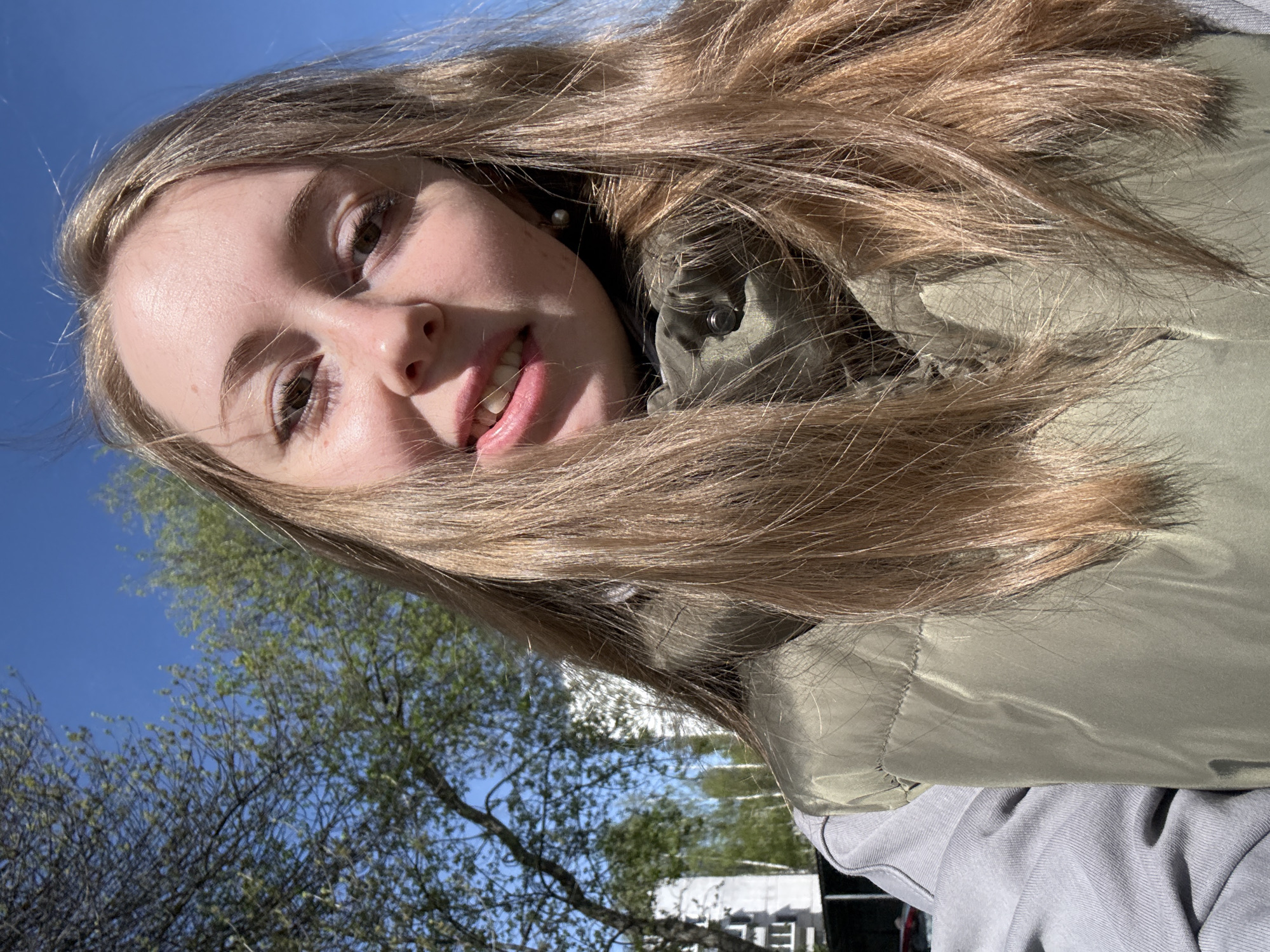 A young woman with long blonde hair, wearing a green padded jacket and grey top, is positioned in a selfie taken outdoors. The image is oriented at an angle with the sky visible behind the subject, along with a tree with budding leaves. The location is the city of Unknown, Unknown.