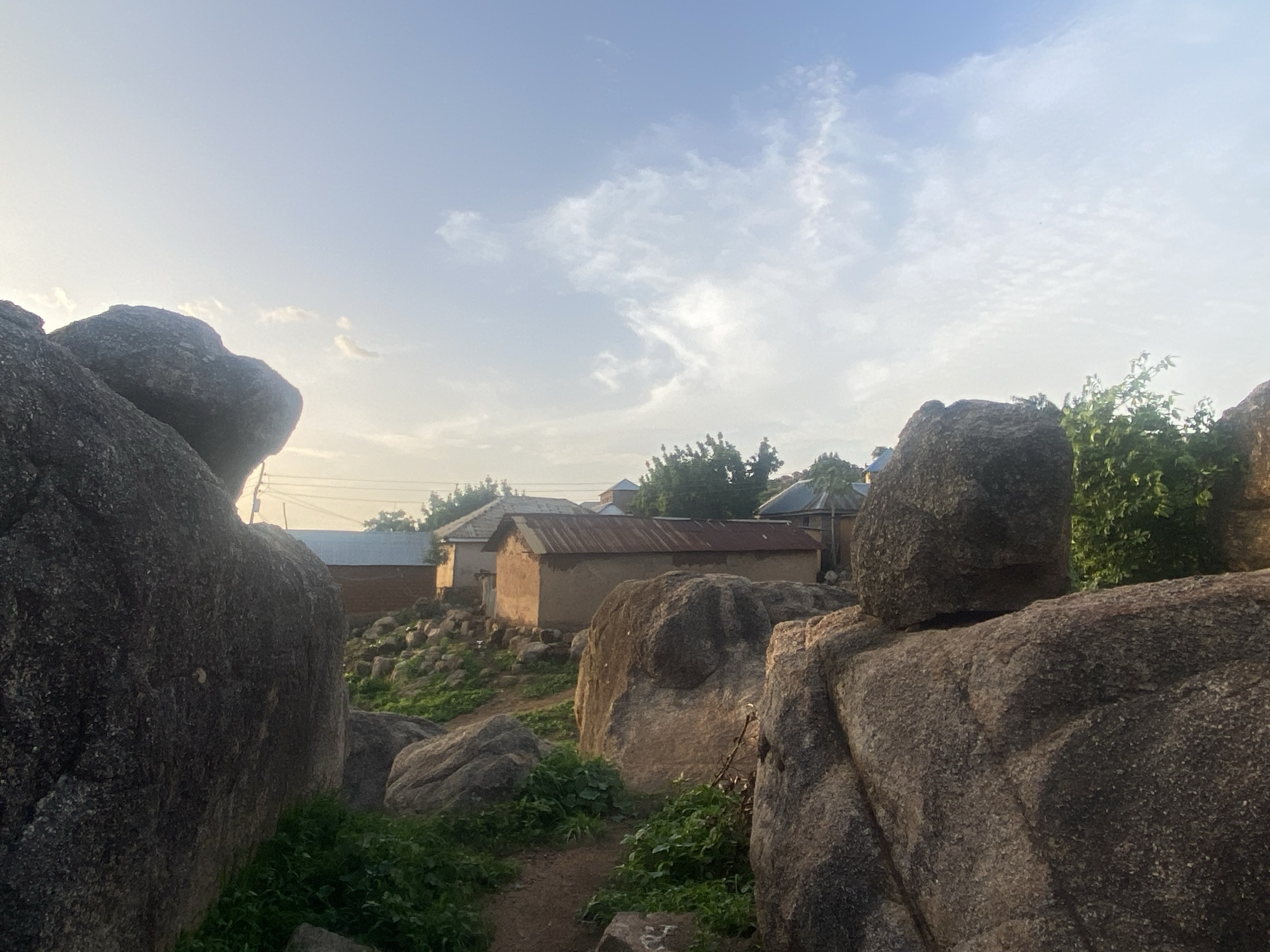 The image shows a rural scene in Jos, Nigeria, featuring large granite boulders in the foreground framing a view of small, mud-walled buildings with corrugated metal roofs. The structures suggest a village or residential area, with lush green vegetation interspersed among the rocks and buildings. The sky is pale blue with scattered clouds, indicating either early morning or late afternoon light. The boulders are gray and weathered, showing varying shades of light and shadow. A narrow, dirt path winds through the rocks, leading toward the buildings. No people are visible in the frame.