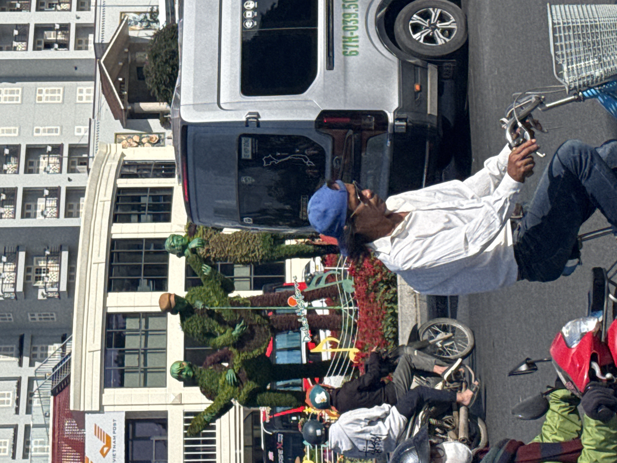 The image depicts a street scene in Da Lat, Vietnam, with a focus on a van and several people on motorbikes. In the background, modern buildings with balconies and windows are visible, suggesting an urban environment. In the foreground, a silver and black van, identified by the license plate "67H-039.50," is parked on the right side of the frame.
Several individuals are present. On the left, two people are seated on a motorbike, one wearing a grey hooded jacket and a dark helmet, the other a black jacket and a light-colored helmet. To the right of them, a man wearing a white shirt, blue cap, and sunglasses is riding a bicycle. Further to the right, part of another person on a motorbike, wearing a green jacket, is visible.
Decorative topiary figures, resembling people, are positioned on the left side of the scene, adding an artistic element to the streetscape. Red flowers and green foliage are arranged around the base of these figures, creating a vibrant display. The overall impression is that of a bustling street in Da Lat, with a mix of everyday life and decorative public art.