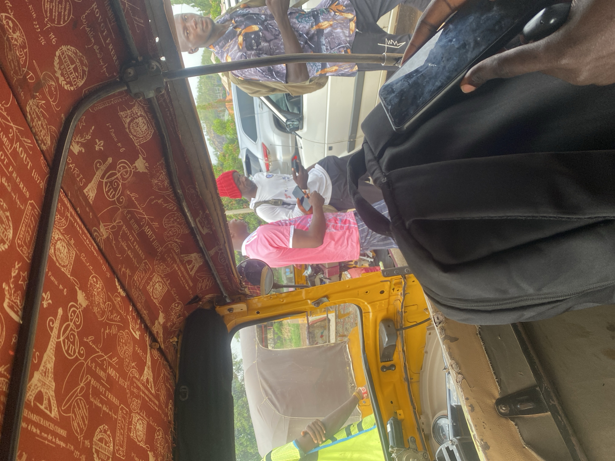 The image shows the interior of a yellow commercial tricycle, commonly known as a "Keke," in Abuja, Nigeria. The interior roof is lined with a red fabric featuring white Parisian themes, including images of the Eiffel Tower and French text. Several individuals are visible outside the Keke, including one in a pink shirt, one in a white shirt with a red cap, and another in a patterned shirt. Passengers appear to be using their mobile phones inside the Keke. A black backpack is also visible inside the tricycle. A car and some greenery can be seen in the background, suggesting an urban environment.