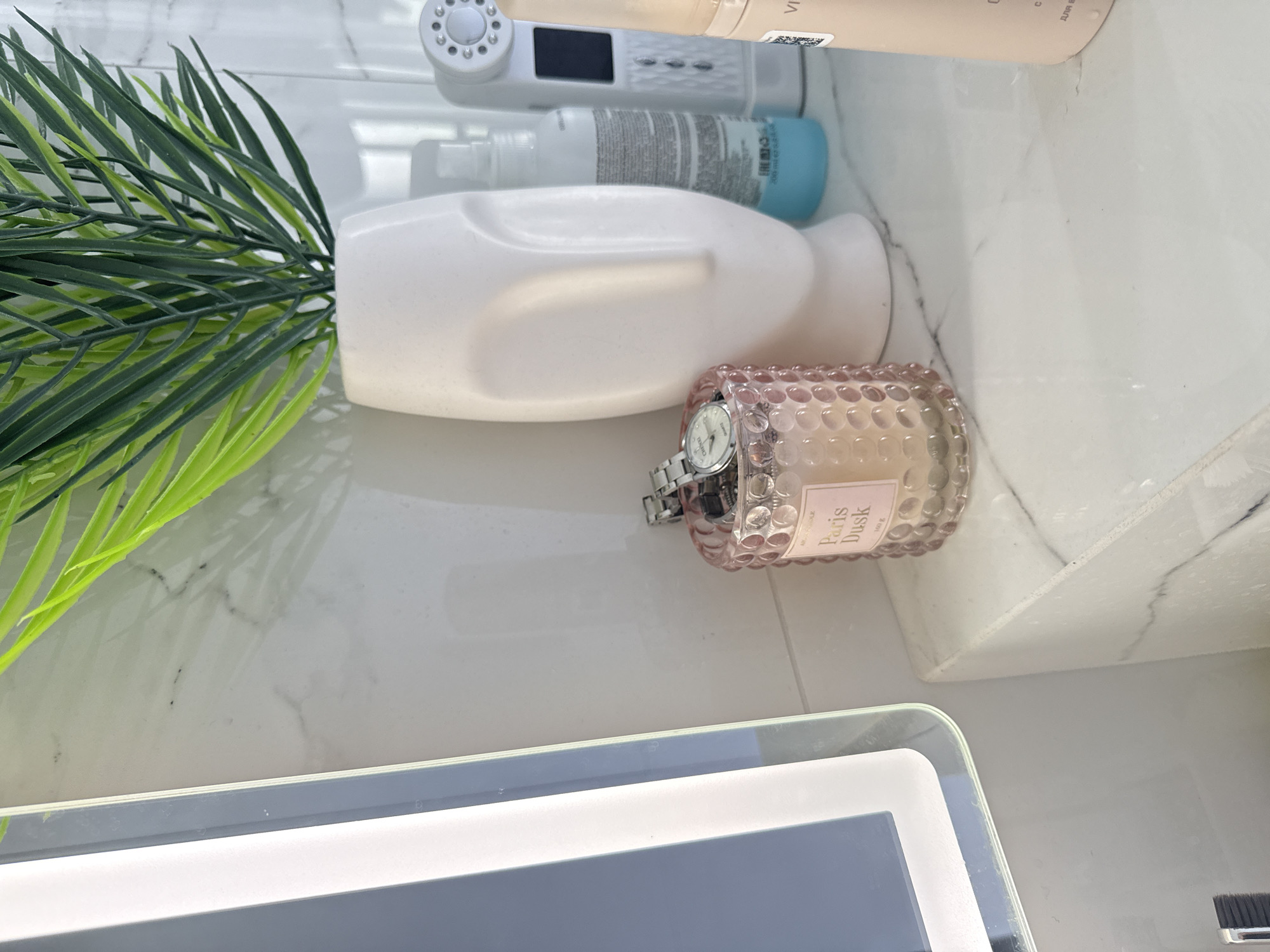 The image shows a close-up of a marble countertop with various items. In the foreground, there is a pink glass candle with the label "Paris Dusk" and a silver watch inside it. To the right of the candle is a white vase with a face design, holding a green artificial plant. Behind the vase are several cosmetic products, including a spray bottle with a blue liquid, a white device with lights and a screen, and a light pink bottle. A large mirror with an illuminated frame is visible on the left side of the image. The background appears to be a tiled wall. The lighting suggests an indoor setting, likely a bathroom or vanity area. The image does not provide any information about the city of Нижнеяркеево, Russia.