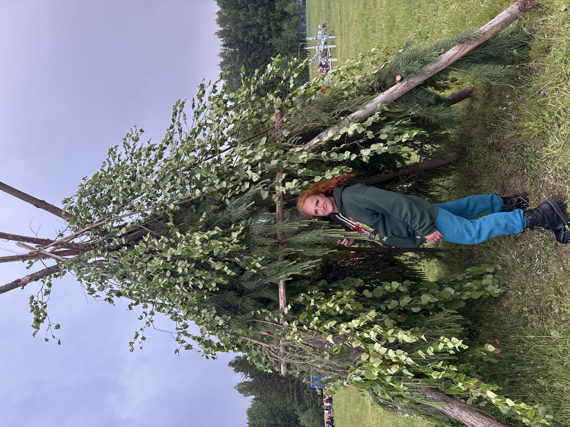 An adult woman with red, curly hair stands in a grassy field in Bol'shaya Kivara, Russia, next to a large, conical, temporary shelter. The woman wears a dark green hooded sweatshirt with a multi-colored graphic, teal pants, and black boots. She is positioned slightly within the shelter's opening, with her left hand touching a branch, and exhibits a faint smile. The shelter is constructed from sturdy tree branches forming a frame, densely covered with both light green deciduous leaves and dark green coniferous boughs. The sky above is overcast and light grey. In the background, a coniferous forest line is visible. To the left, a blurry group of individuals is seated or gathered on the grass. To the right, a collection of white, upright markers, possibly crosses or memorial stakes, are visible in the distance, adorned with various offerings, and a faint plume of smoke rises nearby.