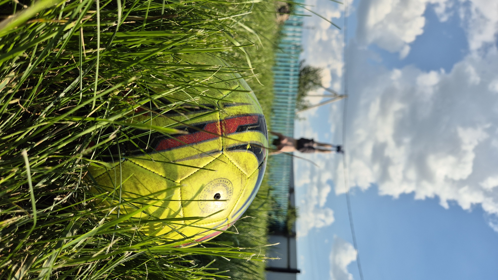A yellow soccer ball with red accents rests partially obscured in tall green grass. In the background, a person is performing a handstand, their legs pointed towards a bright blue sky filled with fluffy white clouds. A light blue wooden fence runs horizontally behind the person, with a few trees and a simple wooden structure visible further in the distance. The scene appears to be outdoors in a rural or suburban setting.