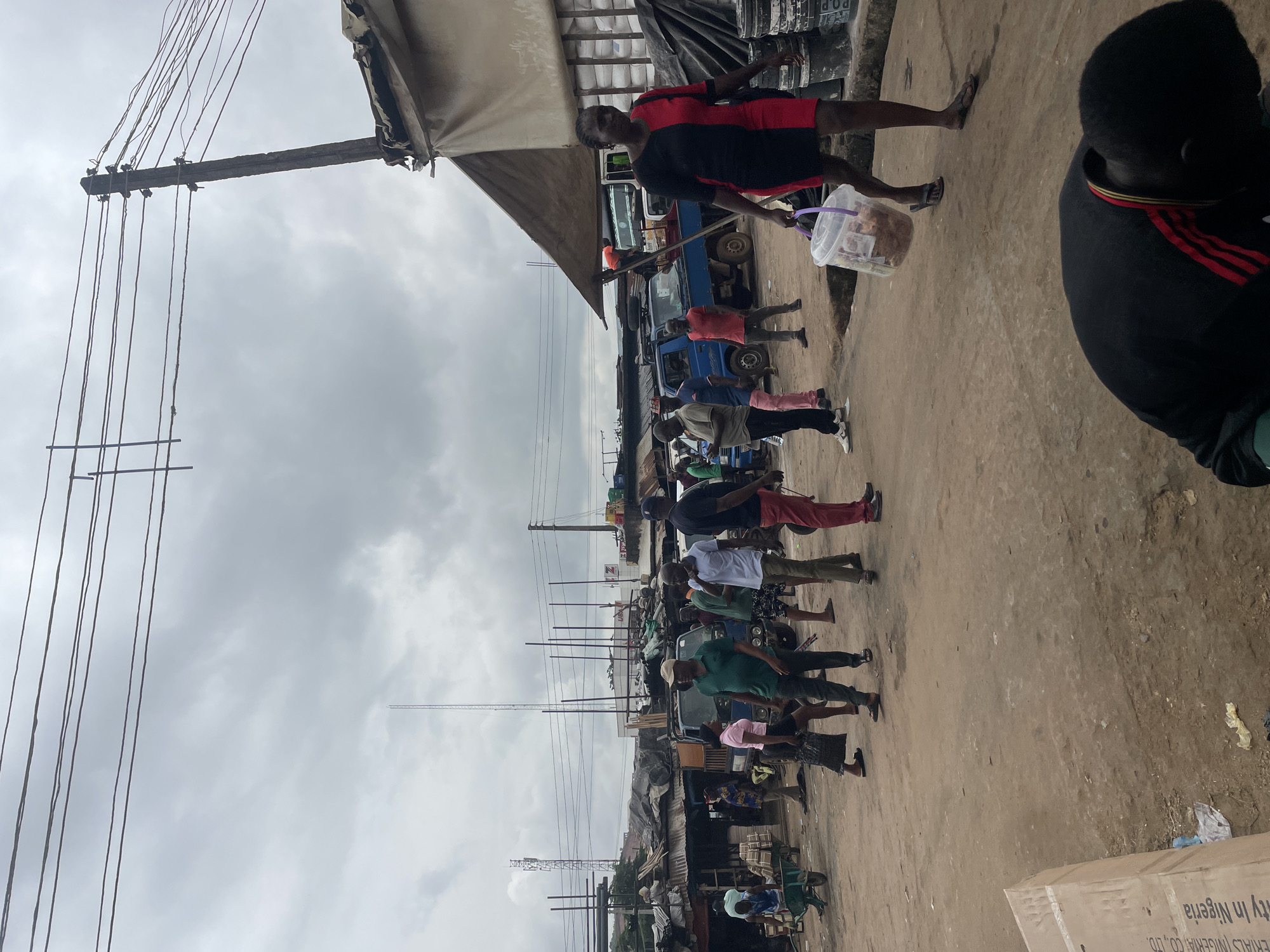 The image depicts a street scene in Ogbunike, Nigeria, under an overcast sky. A variety of people are present, some walking, others appearing to be engaged in activities related to commerce. In the foreground, a person wearing a black shirt with red and yellow stripes is crouched down, partially obscuring the view of a large cardboard box labeled with text in English, including "Materials Nigeria Co., Ltd." and "ity in Nigeria."
Further into the scene, a woman in a two-toned red and black dress walks away from the camera, carrying a clear plastic bucket filled with a light brown substance. Several other individuals are scattered throughout the image. Some are standing and conversing, while others are walking through the dusty, unpaved street. A blue pickup truck is parked in the middle ground, and various stalls or structures with corrugated metal roofing line the sides of the street, suggesting a marketplace or commercial area. Overhead, a network of power lines crisscrosses the sky, supported by a wooden utility pole. A tall transmission tower is visible in the distance. The ground is a mix of dirt and debris.
