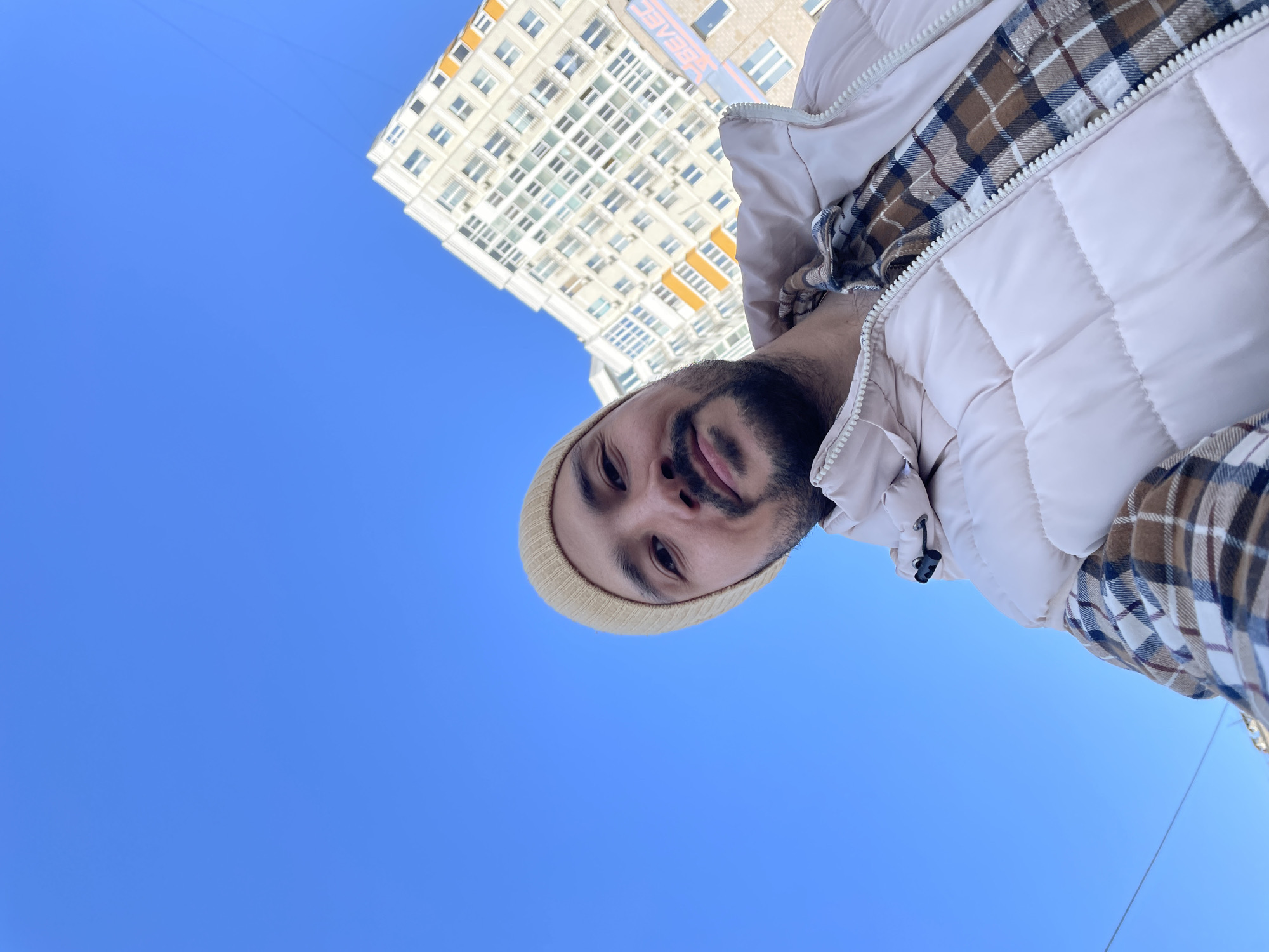 A low-angle selfie depicts a man with dark hair and a beard wearing a tan beanie, a beige puffy vest, and a plaid shirt. The background consists of a clear, cloudless blue sky and a multi-story white building with numerous windows, located in Izhevsk, Russia.