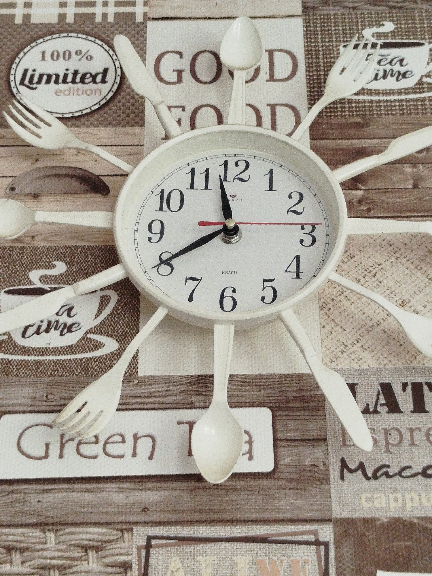 The image shows a close-up of a decorative clock on a textured, wood-grain patterned background. The clock has a white face with black numbers and hands. Surrounding the clock are twelve white cutlery items, including forks, spoons, and knives, arranged radially like the hours on a clock. The background also features various text elements, such as "GOOD FOOD," "100% LIMITED edition," "tea time," "Green Tea," "LATTE," "espre," and "Macc." The overall aesthetic suggests a kitchen or dining area with a culinary theme. There are no people visible in the image. The location "Крывасельскі сельскі Савет, Belarus" is a rural administrative division and does not provide specific information about the visual content of the image itself.