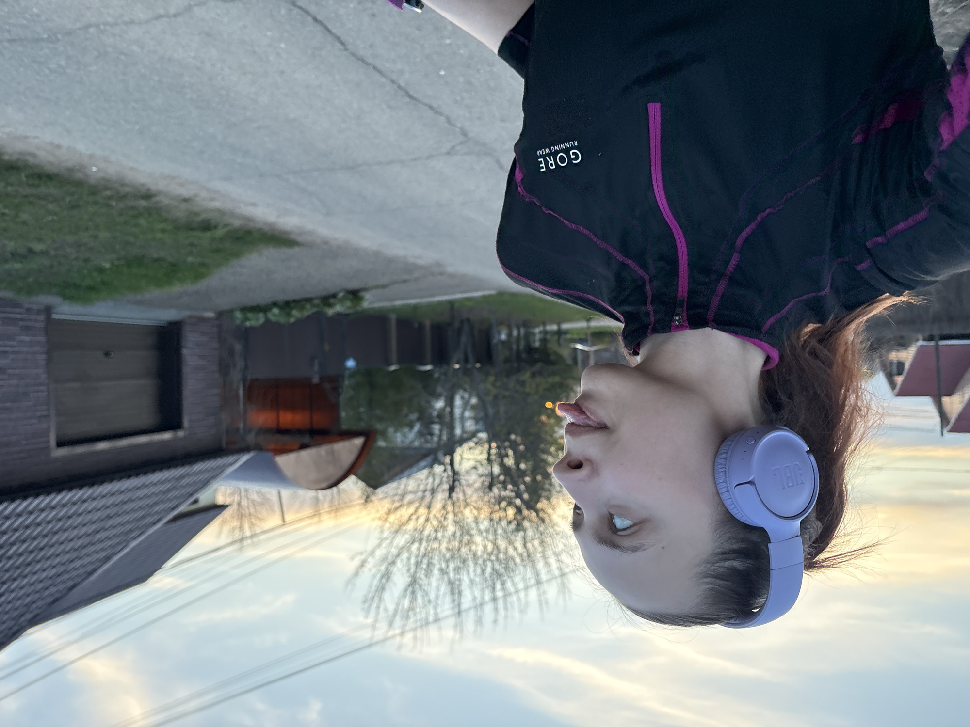 The image shows a woman outdoors in Прикамский, Russia. She is wearing a black Gore running jacket with magenta accents and light purple JBL headphones. The background includes houses with various roof colors, bare trees, and a sky with a sunset or sunrise visible. The woman is standing on a paved area, likely a road or sidewalk.