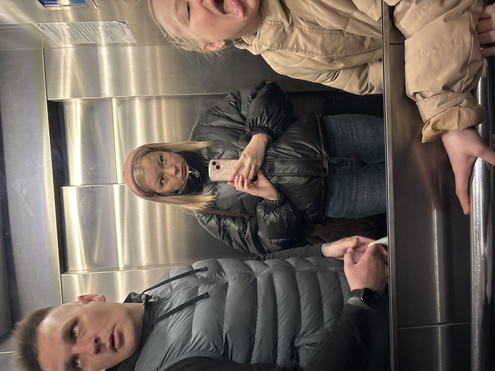 The image shows a group of three people taking a selfie in what appears to be an elevator with stainless steel walls. The location is Izhevsk, Russia.
A man in a grey padded vest and black undershirt is visible at the bottom of the frame, looking towards the camera. Above him is a woman with blonde hair and a pink headband, holding a pink smartphone and looking at the camera. Above her is a young child in a beige jacket sticking their tongue out. The child is also holding onto the metal handrail. The elevator has a panel with information on the top left.