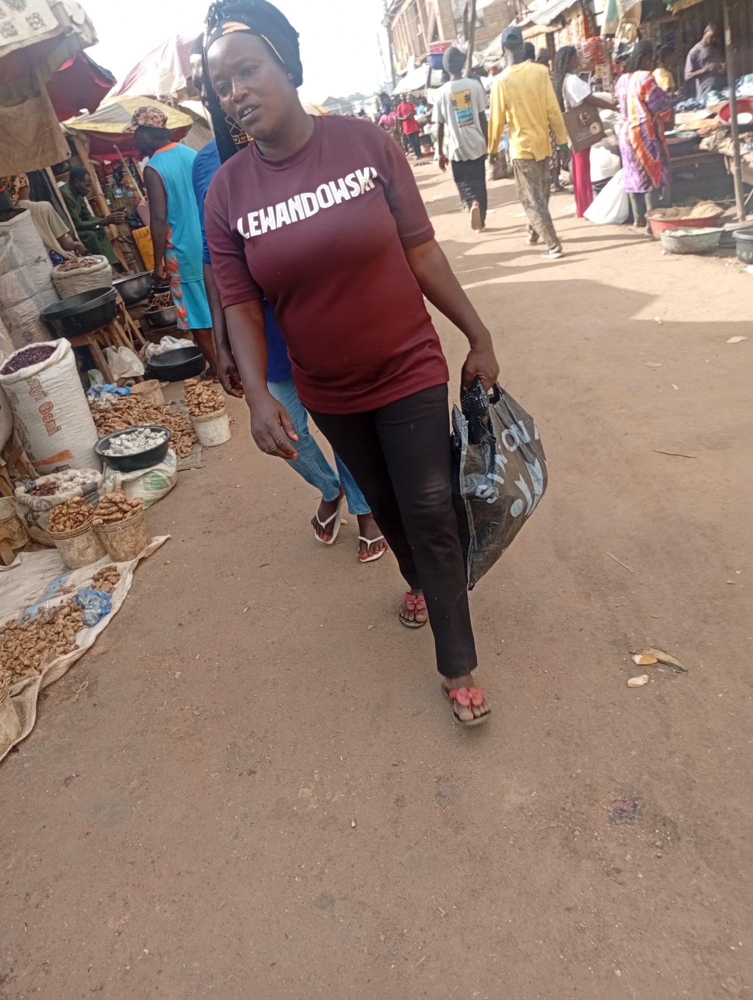 This media file captures a bustling market scene in Rafin Par, Nigeria. The primary subject is a woman walking towards the viewer, carrying a black plastic bag. She is wearing a dark red t-shirt with white lettering and black pants, along with red sandals. Her t-shirt displays the word "LEWANDOWSKI" in prominent white letters.
Behind her, other individuals are present, some walking away from the camera and others tending to market stalls. To the left of the frame, several stalls are visible, laden with various goods. These include sacks of what appear to be grains or beans, baskets filled with ginger or similar root vegetables, and open containers with produce. The ground is a dusty, reddish-brown earth, characteristic of the region.
The lighting suggests it is daytime, likely mid-morning or afternoon, with the sun casting shadows and illuminating the scene. The atmosphere appears active, typical of a local market where commerce and daily life intersect. The overall impression is one of a vibrant and busy marketplace.