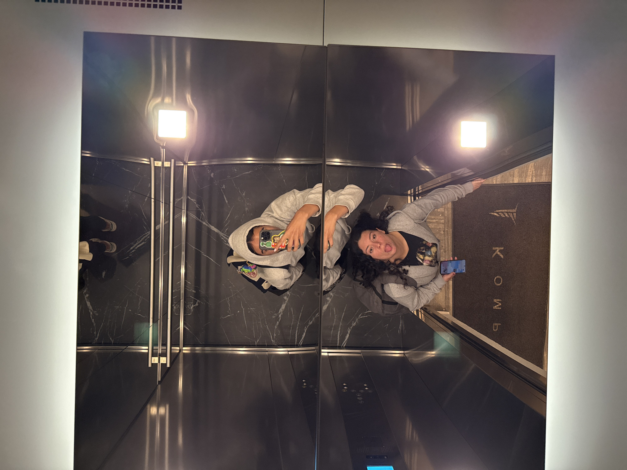 Two young women take a selfie in a mirrored elevator. One woman is wearing a gray hooded sweatshirt and holding a cell phone up to take the photo. The other woman is wearing a gray hooded sweatshirt as well and sticking her tongue out. The elevator has a black marble design. A light fixture is visible at the top. The floor has a mat with the word "KOMB" written on it. The elevator is likely located in the city of Puebla, Mexico.
