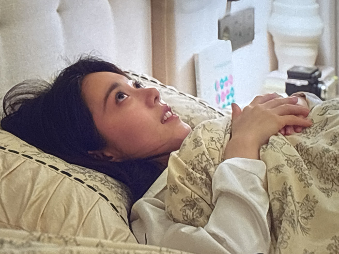 This scene is located in a room in the city of Howrah, India. A female individual, appearing to be of East Asian descent with long, dark hair, is lying reclined on her back in a bed. Her head rests on a light-colored pillow with dark embroidered lines, turned slightly to her right, and her gaze is directed upwards and to her left with a neutral expression and slightly parted lips. She is covered from the chest down by a light-colored blanket or duvet featuring a dark floral or toile-like pattern. Her hands are clasped over the blanket on her chest. A white long-sleeved garment is visible beneath the blanket on her left arm. The background includes a light-colored tufted headboard behind her and, to her right, a white fluted decorative object or lamp base, a partially visible book or framed item with alternating pink and blue oval shapes, a small dark electronic device, and a small rectangular object hanging on the wall.