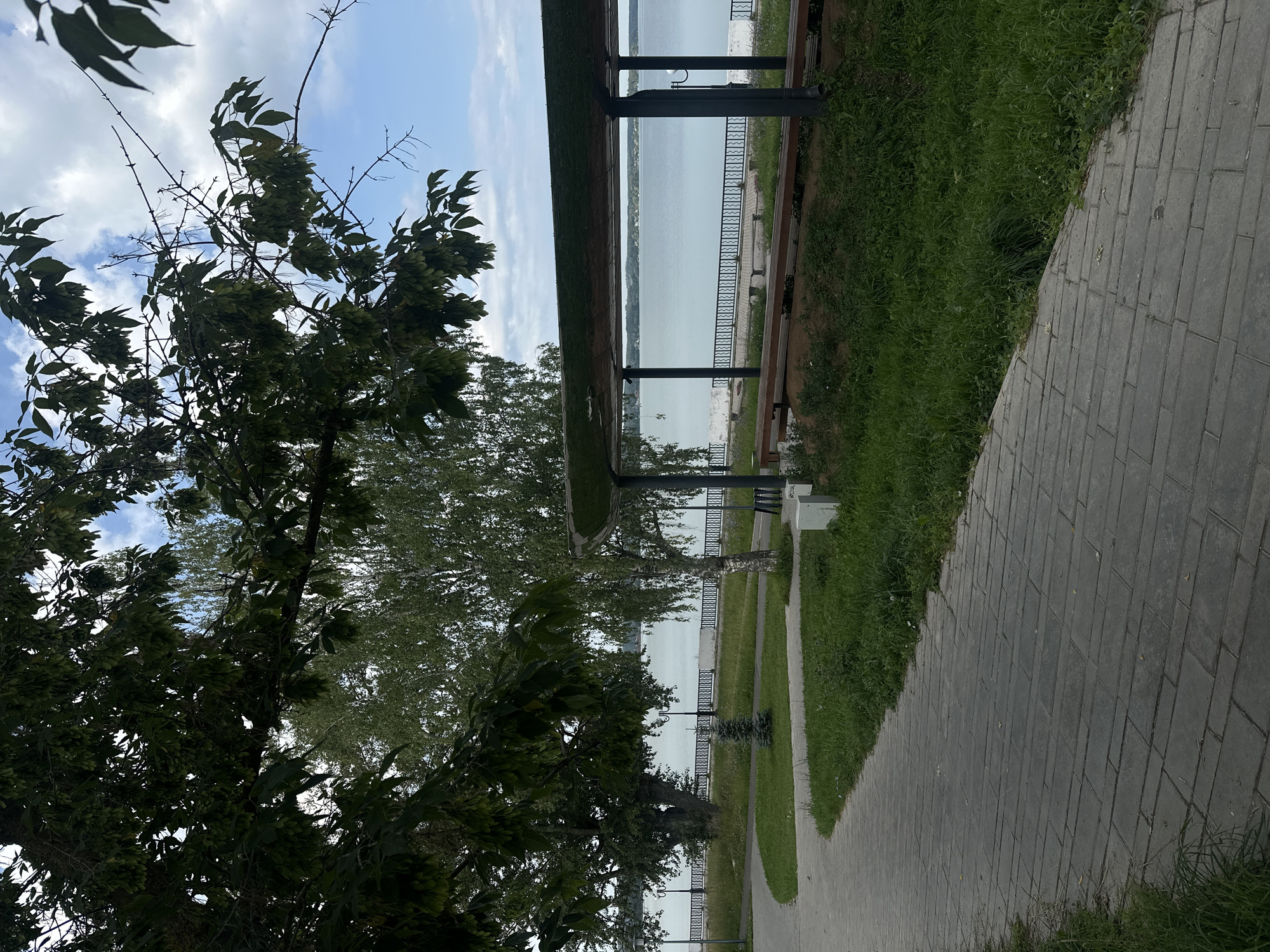The image depicts a scenic waterfront promenade in Votkinsk, Russia. A paved walkway curves through a green grassy area, leading towards a large body of water. Mature trees with dense foliage line the left side of the path, their branches extending into the frame. In the background, a light-colored building with a dark, angled roof is visible, providing shade over a wooden bench. Beyond the structure, a calm, pale blue lake stretches to the horizon, dotted with distant structures. A black metal fence runs along the lake's edge, separating the park area from the water. The sky above is a mix of blue and white clouds. The scene is tranquil and suggests a public park or recreational area. There are no people visible in the image.