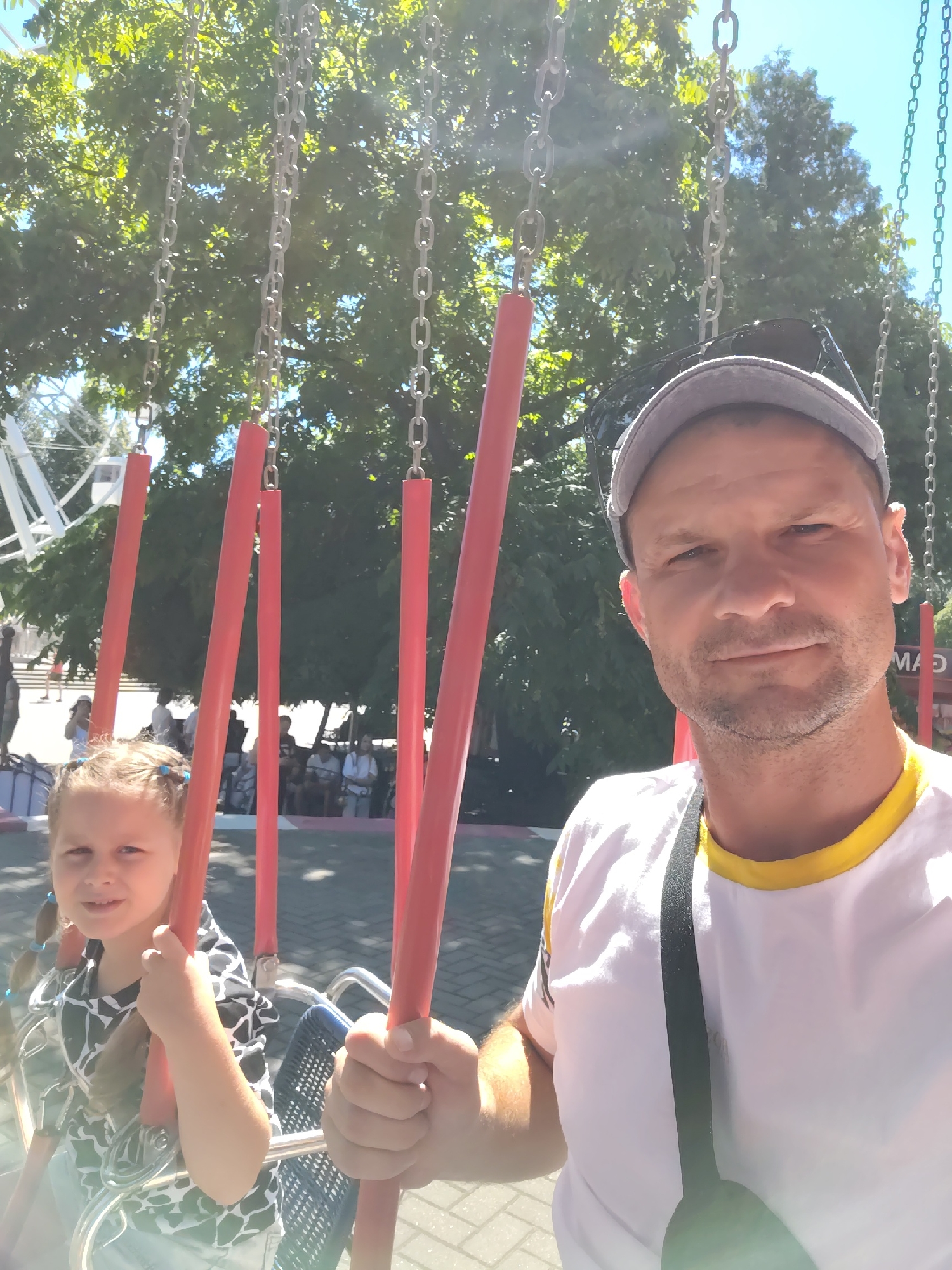 In a selfie taken outdoors, a man wearing a grey baseball cap and a white t-shirt with a yellow trim smiles as he holds onto the frame of a swing ride. Next to him, on his left, a young girl with pigtails and blue hair ties smiles and holds onto the red bars of the swing. Both are seated on a swing in what appears to be an amusement park. In the background, lush green trees, a Ferris wheel, and several other people can be seen. The image is bathed in sunlight, suggesting it was taken on a bright day. The overall setting indicates a recreational outing in a park environment.