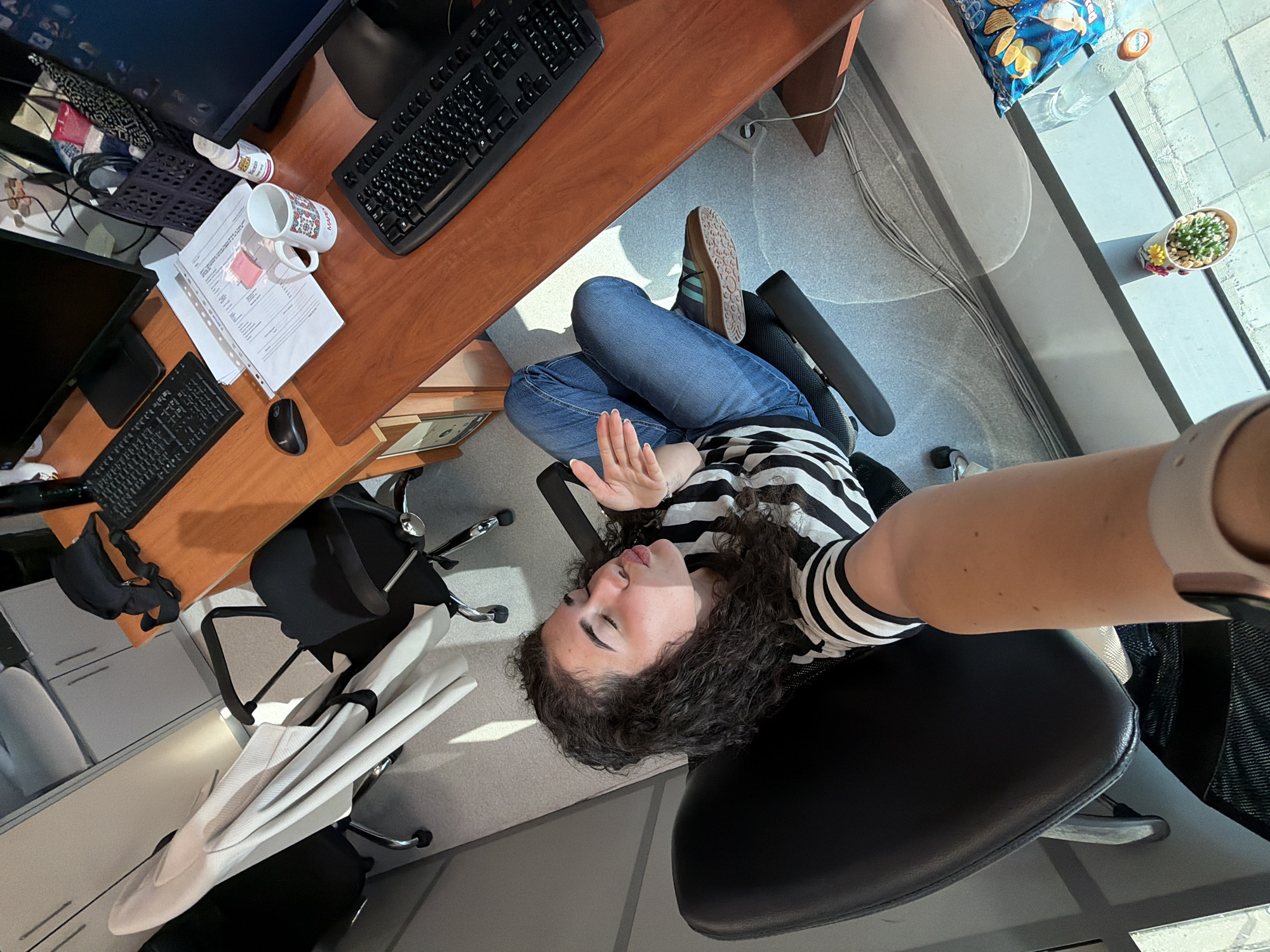 A young woman with curly dark hair is sitting in a black office chair, facing a wooden desk with two computer monitors. She is wearing a black and white striped shirt and blue jeans, and has her eyes closed with her lips pursed as if blowing a kiss. Her right arm is extended towards the camera, holding a smartphone in a selfie-taking position. The desk is cluttered with office supplies, including keyboards, a mouse, papers, a mug, and a bottle of pills. A basket filled with snacks sits to the right of the monitors. In the background, two other office chairs are visible, one with a light-colored garment draped over it. The scene appears to be an office environment, likely in Sofia, Bulgaria, given the detail. A plant in a small pot and a bag of chips are on the windowsill in the foreground, suggesting the office might be located on an upper floor with a view of an outdoor area.
