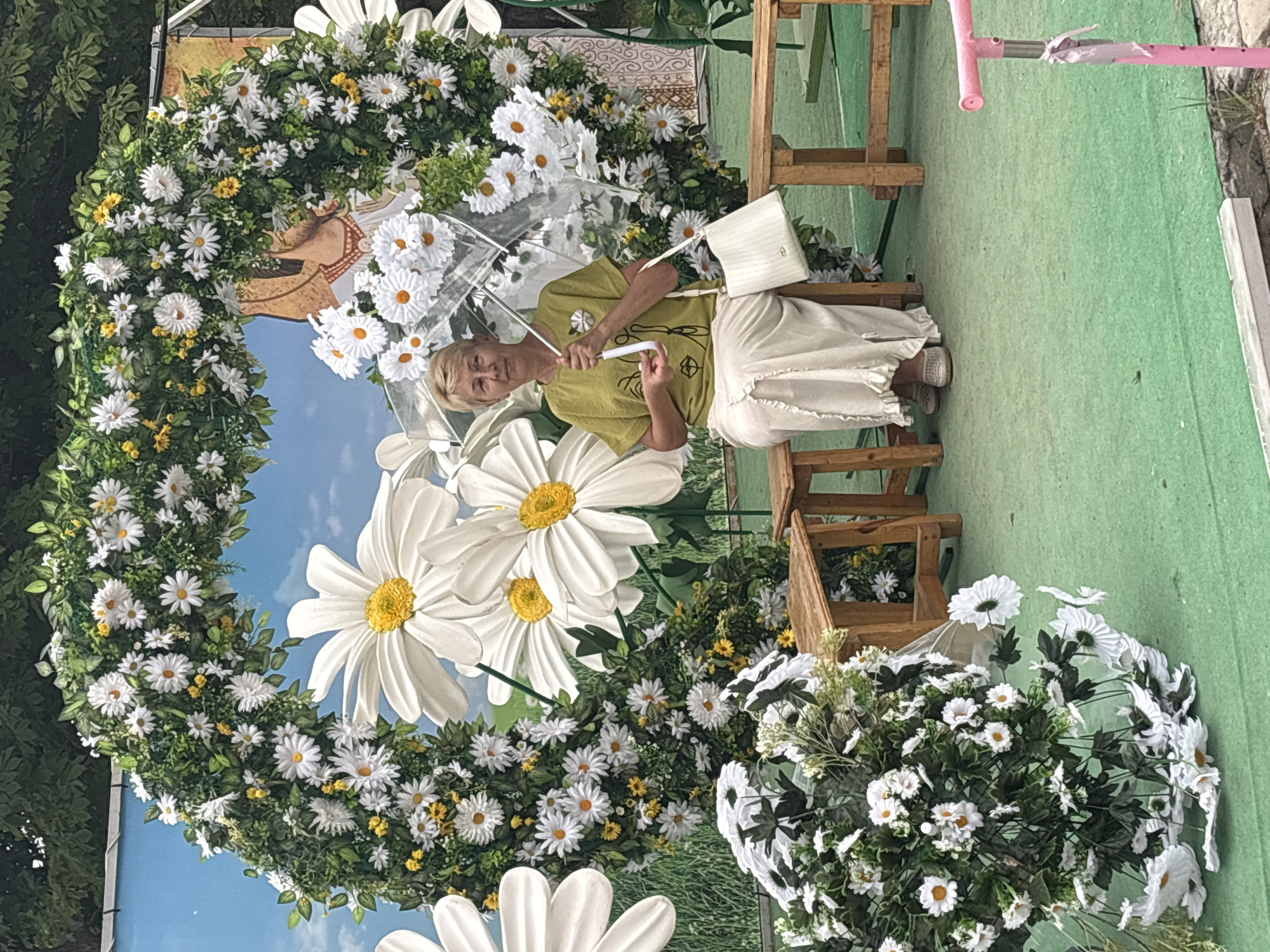 A middle-aged woman with short blonde hair is sitting on a wooden bench, holding a clear umbrella with daisies. She is wearing a short-sleeved olive green t-shirt with a graphic print of a face and a long, flowing cream-colored skirt. Next to her is a white handbag. She is seated in front of a large floral archway adorned with white daisies and greenery, with a backdrop depicting a blue sky and grass. The scene appears to be outdoors, possibly a park or a decorative setup for an event. In the foreground, to the left, is a large bouquet of white flowers. Wooden benches are also visible to her right. The overall setting suggests a festive or decorative outdoor environment, potentially related to a local celebration or public event in Astrakhan, Russia.