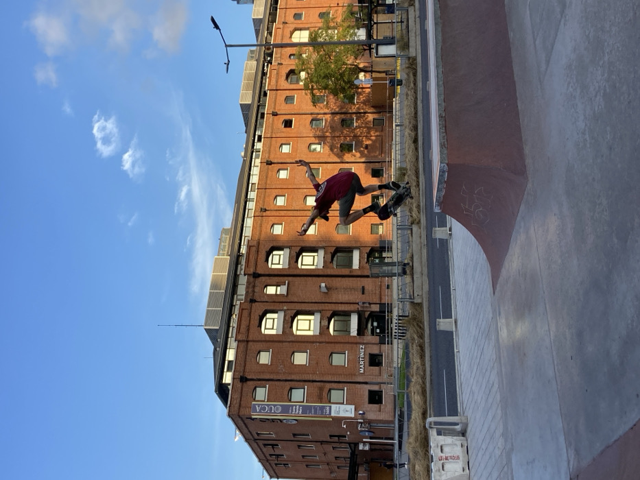 A skateboarder mid-air, performing a trick near a skate park edge. The background features a multi-story brick building with windows, signs, and possibly solar panels on the roof. A street lamp stands nearby. The sky is blue with some clouds. The image orientation is vertical.