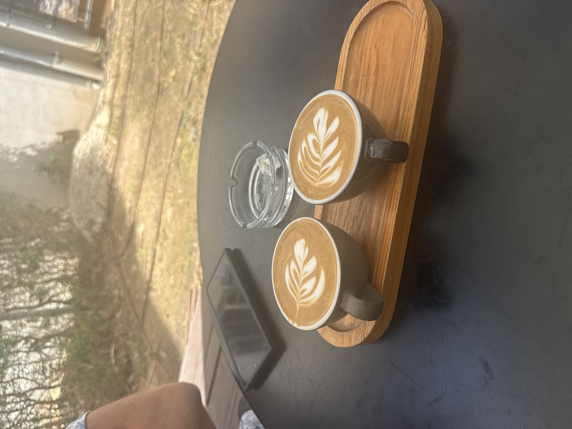 The image depicts a table setting outdoors, featuring two cups of latte art coffee on a wooden tray. Beside the tray sits a glass ashtray and a black smartphone. The table has a dark, matte surface. In the blurred background, there is vegetation, possibly trees and grass, and a building structure. Based on the location information, this scene is likely situated in Tel-Aviv, Israel, potentially in a cafe or outdoor seating area. There are no people visible except for a small portion of a person's leg and clothing.