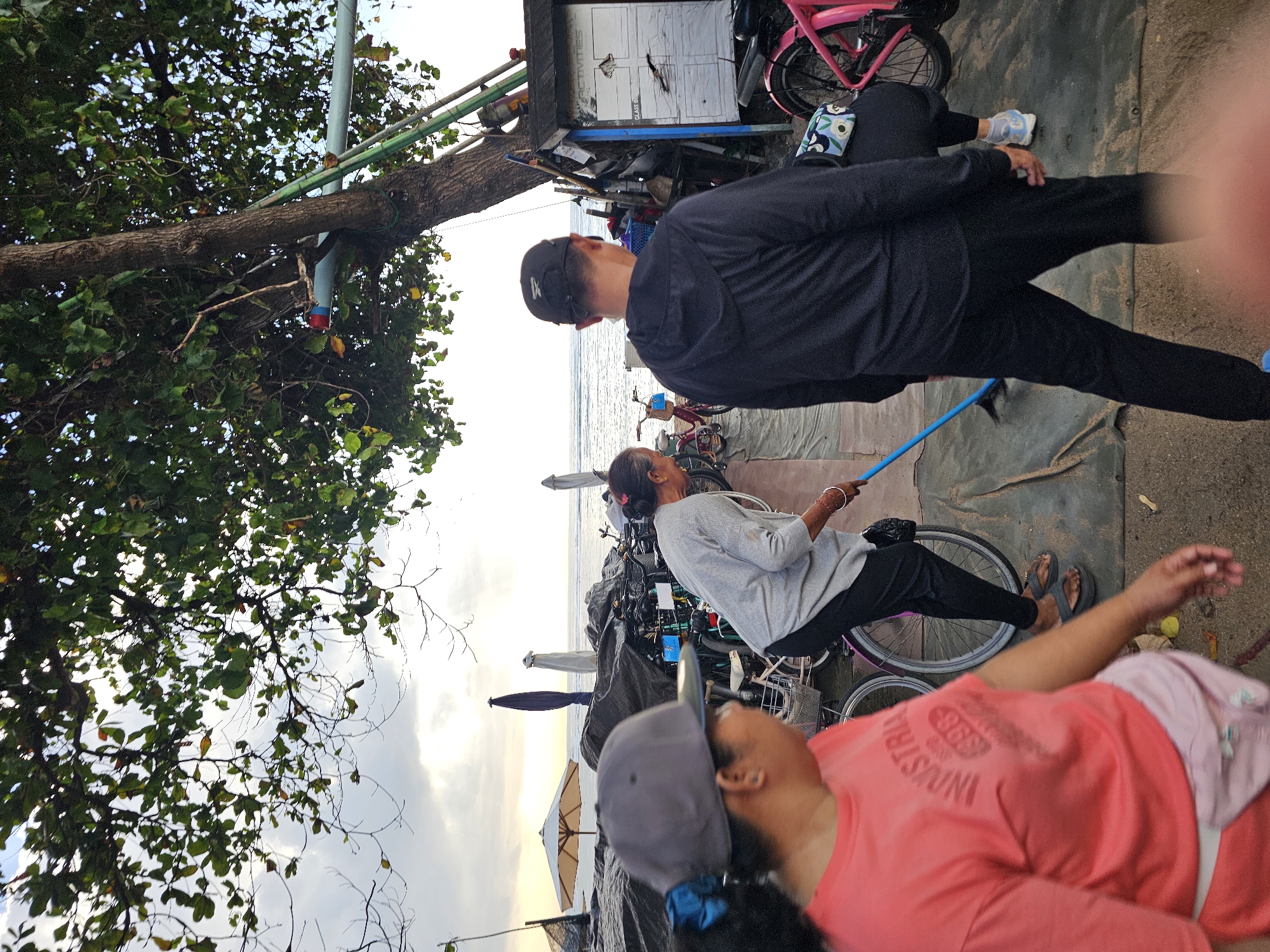 At an outdoor location near the ocean, possibly a beach or promenade in Denpasar, Indonesia, three individuals are present. In the foreground, a person wearing a grey baseball cap and a coral-colored t-shirt with "INDUSTRI 1998" printed on it, is walking away from the camera. Their left hand is visible, and they are wearing a light pink fanny pack.
To their right, a woman with grey hair, wearing a light gray long-sleeved shirt and black pants, is actively sweeping the ground with a blue-handled broom. She is wearing dark flip-flops and has a small black bag slung over her shoulder.
Behind the sweeping woman and to the right, a man wearing a black hooded jacket and black pants is walking away from the camera. He has a black baseball cap on.
The environment appears to be a somewhat cluttered area with several bicycles parked or stored behind the people. Some of the bicycles are pink, and one is white. Tarps and dark materials are spread on the ground, suggesting storage or a temporary setup. In the background, the ocean is visible with waves, and several beach umbrellas and upright poles are present. A tree with dense green foliage extends over the scene from the right. A small structure resembling a shed or booth with white siding and a sign that says "ACTIVITIES" is also visible near the tree. The sky is overcast with some sunlight breaking through.