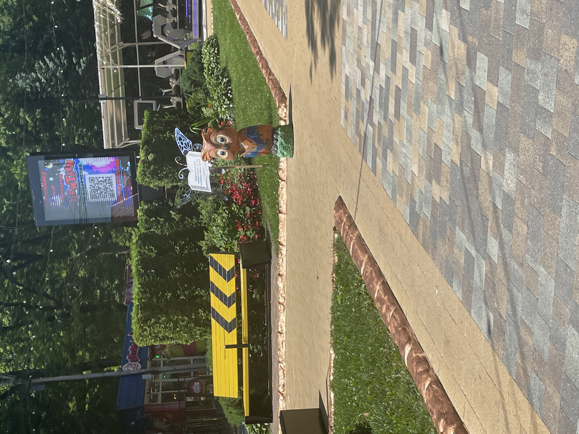 A brightly lit outdoor scene, likely a park or public recreational area, displays a vibrant yellow bench with black chevron markings, situated beside a manicured green hedge. In the foreground, a whimsical owl sculpture, adorned with blue butterfly wings, stands on a green mound. Adjacent to the path, a detailed brick-like border lines a grassy area. Further back, a large digital billboard showcases a QR code and text in Russian, accompanied by an indoor gym facility with exercise machines. The overall environment suggests a lively and well-maintained public space.