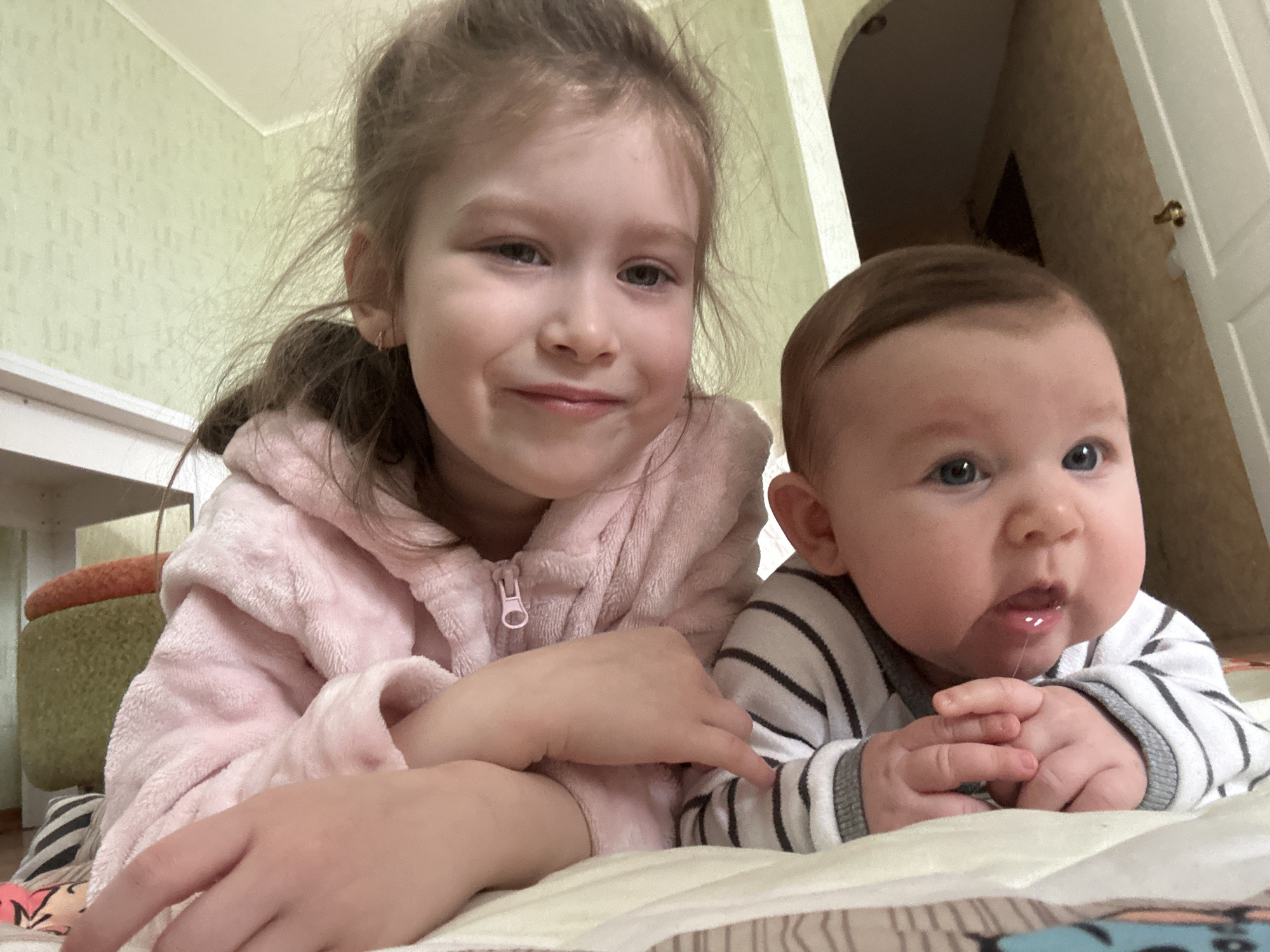 Two children are lying on a patterned surface indoors. The older child, a girl, is wearing a pink hooded garment and has light brown hair pulled back. She is smiling. The younger child, an infant, is wearing a striped top and is drooling. In the background, there is a wall with light-colored wallpaper. Part of a white desk is visible on the left. Further back, there is a doorway or archway. The room appears to be in a residential setting. The location is Chaikovsky, Russia.