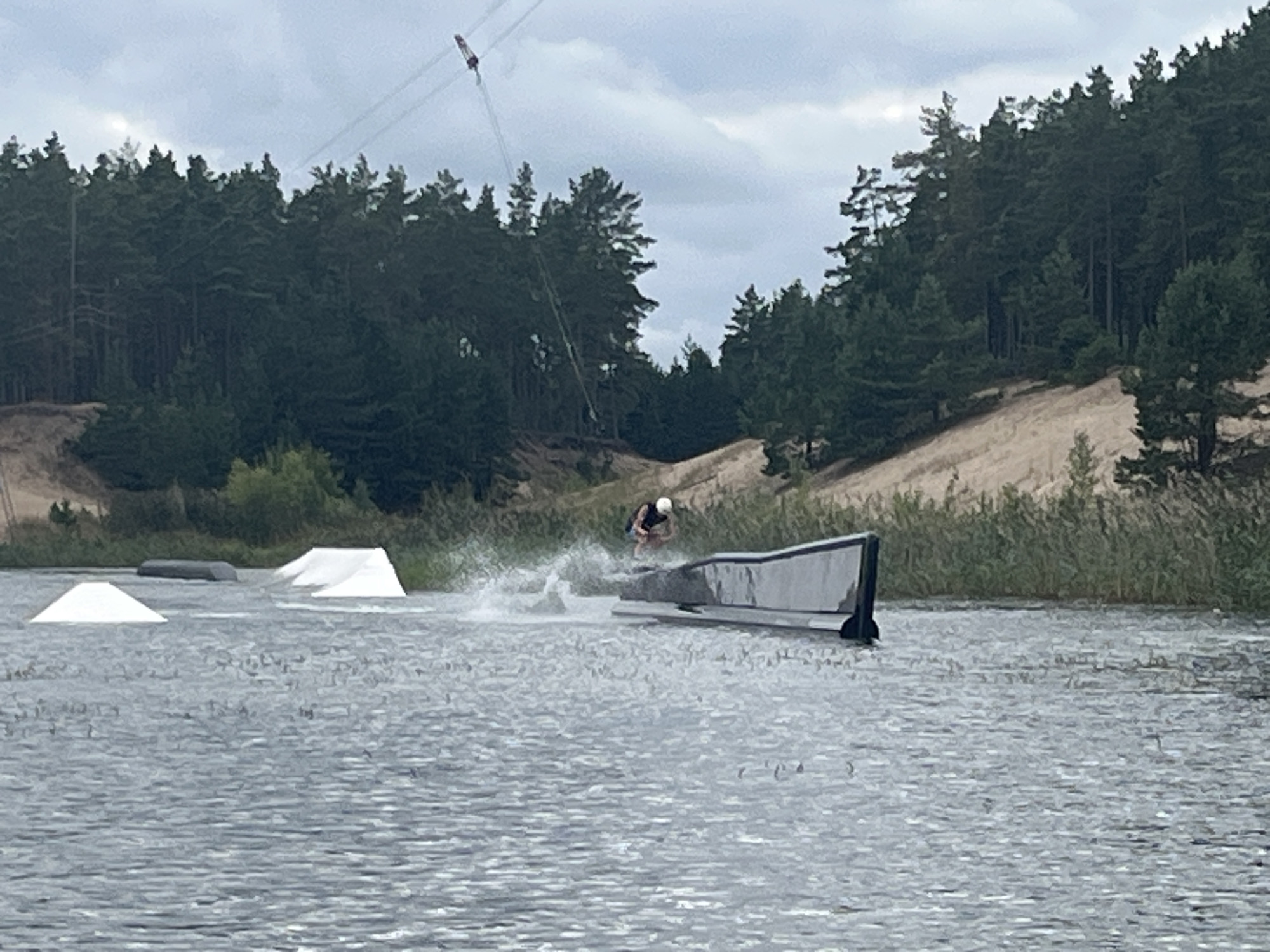 In Männiku, Estonia, a wakeboarder is captured mid-air, performing a trick over a dark, rectangular obstacle in a lake. The wakeboarder, wearing a helmet and dark shorts, is kicking up a significant spray of water. Several white ramps are visible in the water, with one to the left and another further back to the right. In the background, a dense forest of dark green coniferous trees lines a sandy incline. Overhead, a cable system is suspended between two points, with a small red and white object attached to it, likely part of the wakeboard park's infrastructure. The sky is overcast with a light gray cloud cover.