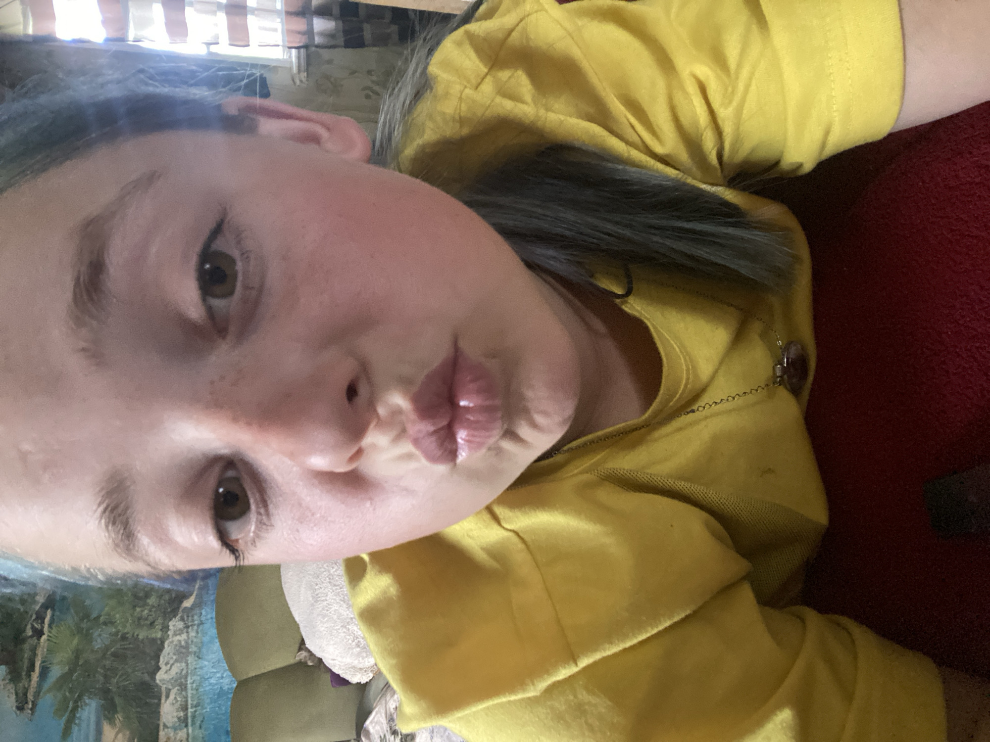 A young person with blue-tinted hair and freckles is taking a selfie indoors. They are wearing a yellow t-shirt and have their lips pursed for a kiss. The background shows a mural of a tropical scene with palm trees and water, and a window with blinds. There are also hints of furniture, including a green sofa and a white pillow. The overall impression is of a casual, personal moment captured in a home environment. There is no indication of the specific location within Nova Praha, Ukraine.
