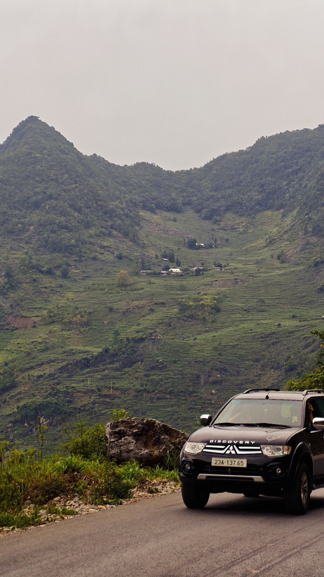An eye-level, medium shot depicts a dark-colored Mitsubishi SUV parked on a paved road in Cán Tỷ Commune, Vietnam, set against a backdrop of verdant, terraced hills and a cloudy sky. The SUV is positioned to the right, with its license plate, “23A-137.65,” visible. A large rock and some shrubbery appear on the roadside beside the vehicle. The terraced hills are densely covered with vegetation, and a small cluster of buildings is visible in the distance. The sky is overcast, casting a muted light over the scene.