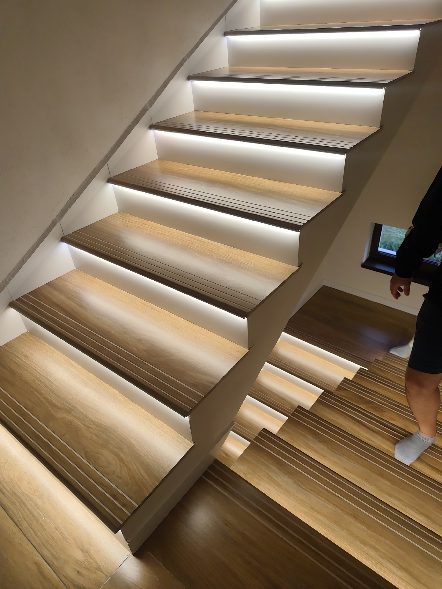 The image depicts a well-lit staircase in an indoor setting. The stairs are constructed with wooden treads and white risers, with integrated LED lighting underneath each tread. The wooden treads feature a grooved design. A person wearing shorts and socks is partially visible on the lower steps, descending the staircase. A small window is present on the right side of the image near the stairs. The location is Mosomishte, Bulgaria.