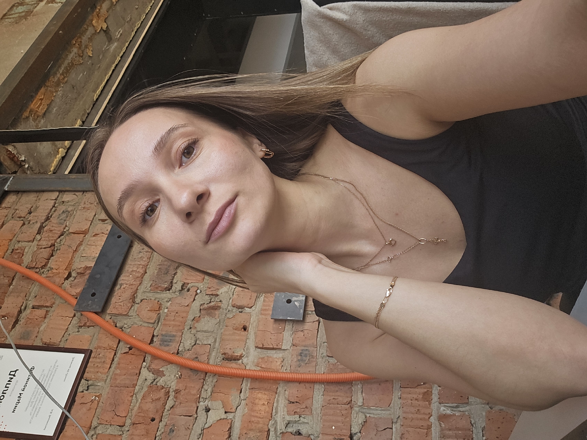 A woman with light brown hair and fair skin is posing for a selfie. She is wearing a black tank top and gold jewelry, including a necklace, earrings, and a bracelet. Her left hand is resting on her neck. The background consists of a red brick wall with an orange conduit running along it, a certificate or sign in the lower left corner, and what appears to be exposed ceiling beams and framing. The photograph was taken indoors in Chaikovsky, Russia.