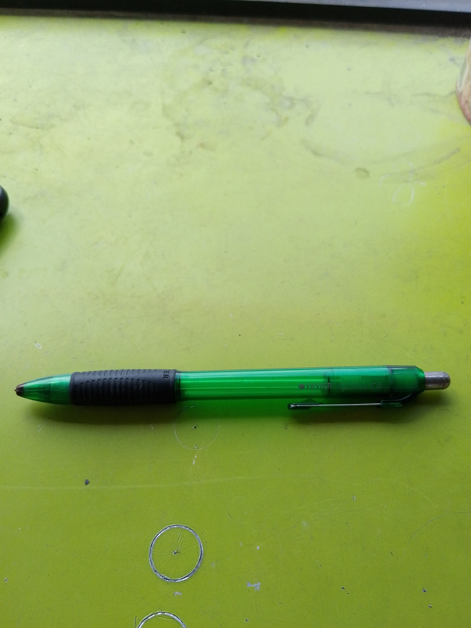A green retractable pen with a black grip is lying horizontally on a bright green, textured surface. The pen is positioned in the lower half of the image, extending from the left to the right. The grip is ridged and appears to be made of rubber. A silver clip is attached to the pen body near the tip. On the green surface, there are several faint, circular indentations or marks, some of which appear to be rings. The background is out of focus, showing a darker surface at the top. The overall scene is a close-up view of the pen and the surface it rests on. No specific location in Surabaya, Indonesia, can be determined from the image.