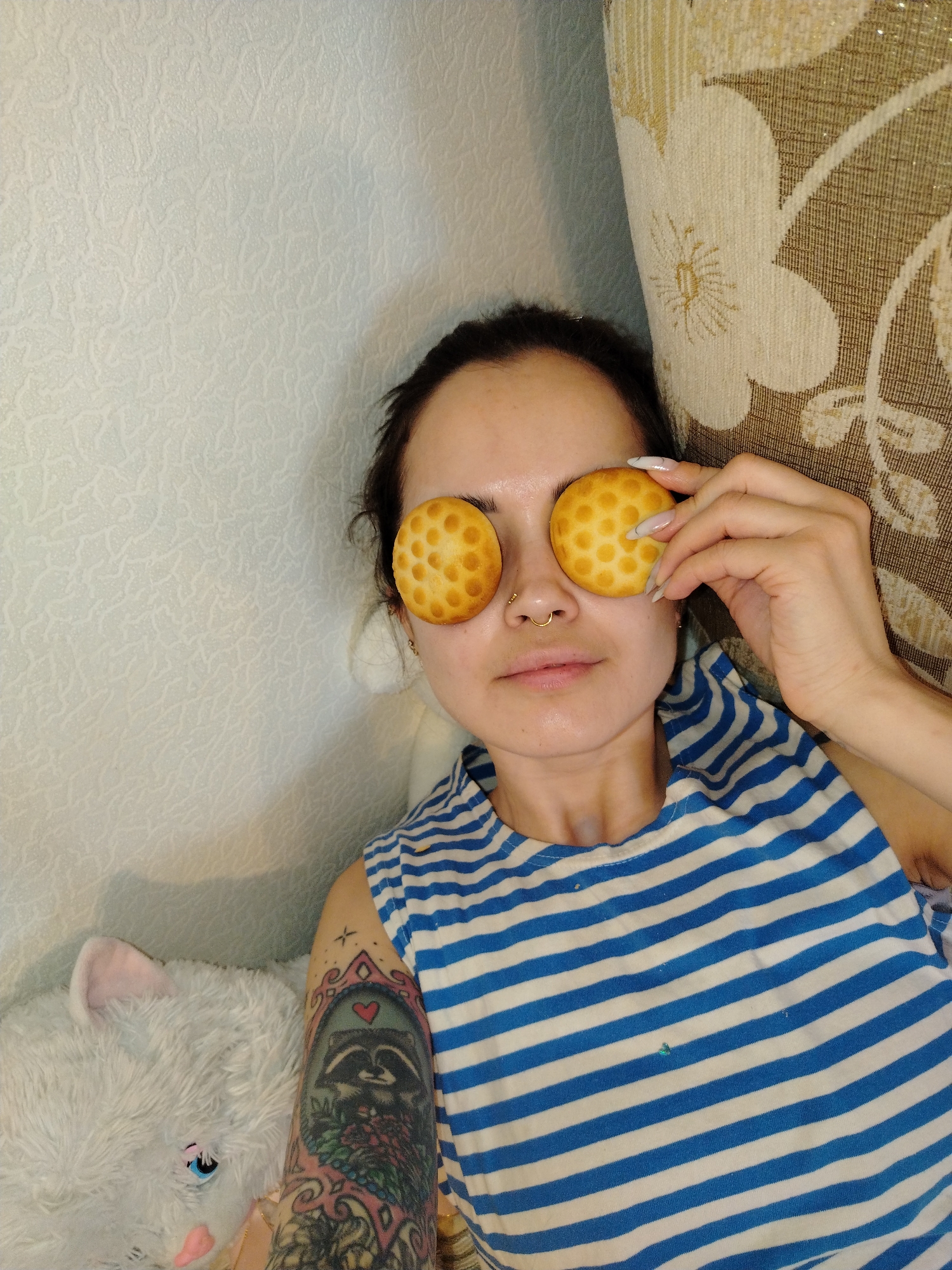 A woman with dark hair pulled back is lying down, holding two round, dotted cookies over her eyes. She has a nose ring and is wearing a blue and white striped shirt. Her left arm, visible on the lower left side of the image, has a tattoo of a raccoon. A fluffy white stuffed animal is positioned next to her. The background includes a textured off-white wall on the left and a floral patterned pillow on the right. The city and location are unknown.